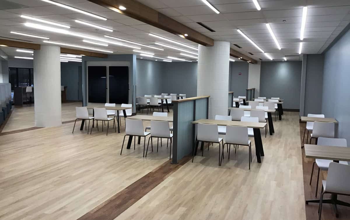 Open dining room with rows of wood tables and white chairs beneath linear LED lights.