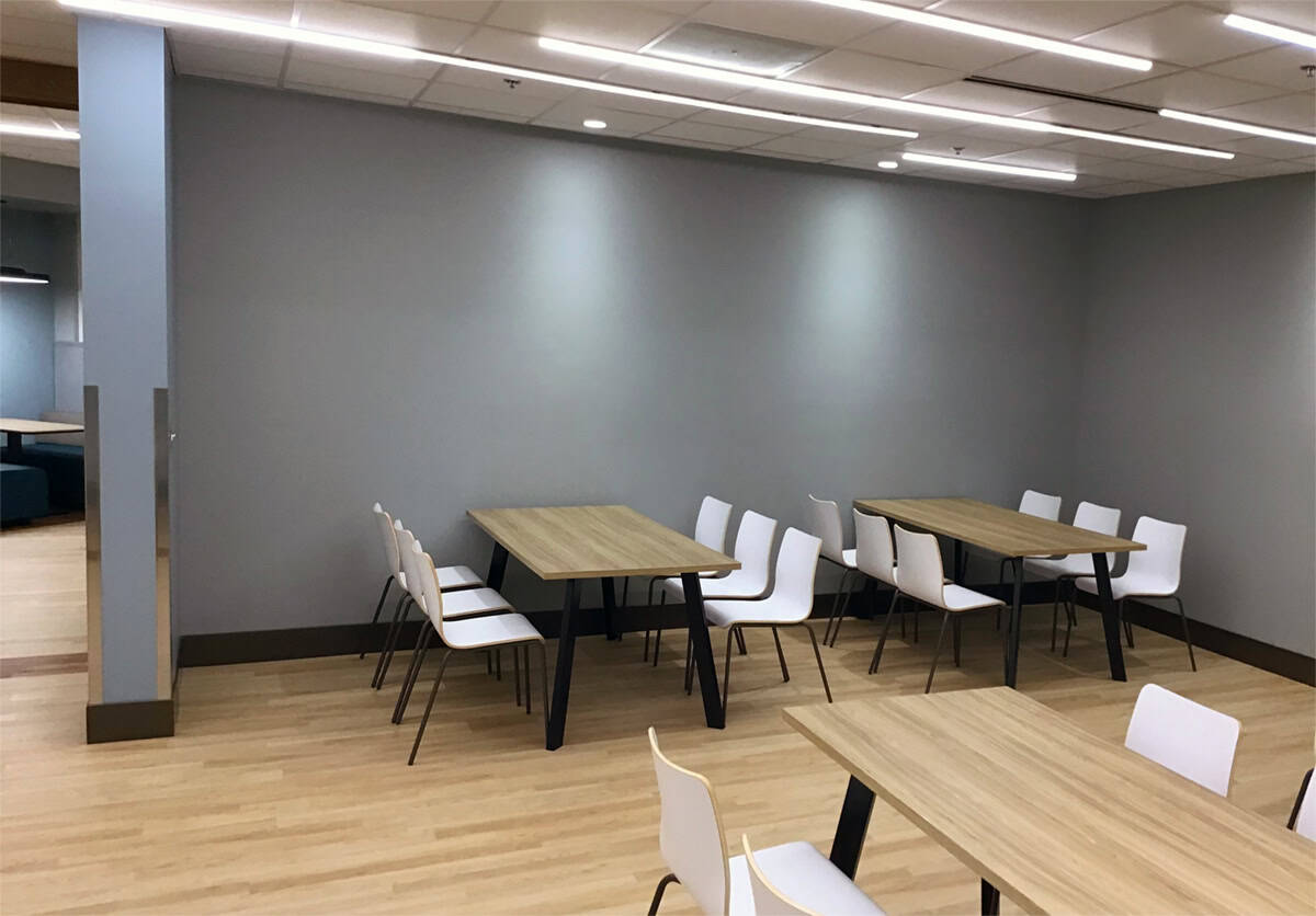 Wood dining tables and white chairs against a gray-blue wall in the hospital cafeteria.