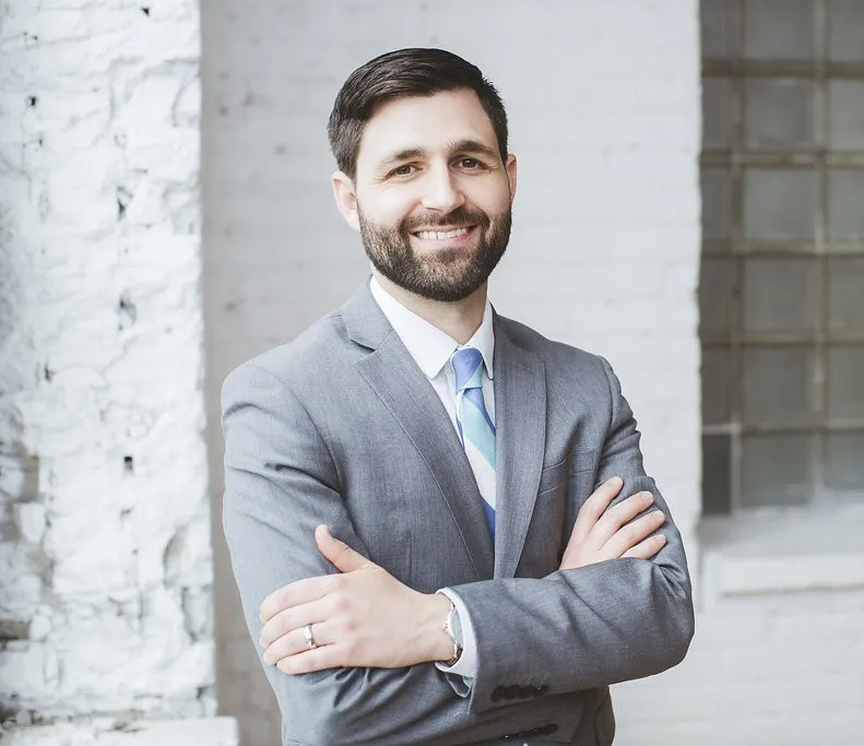 Dr. David Bates standing confidently in a gray suit with arms crossed, smiling in front of a white brick wall background.