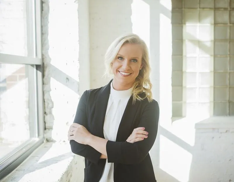 Dr. Katie Hannon smiling with arms crossed in a sunlit room, dressed in business attire with a confident and welcoming posture.