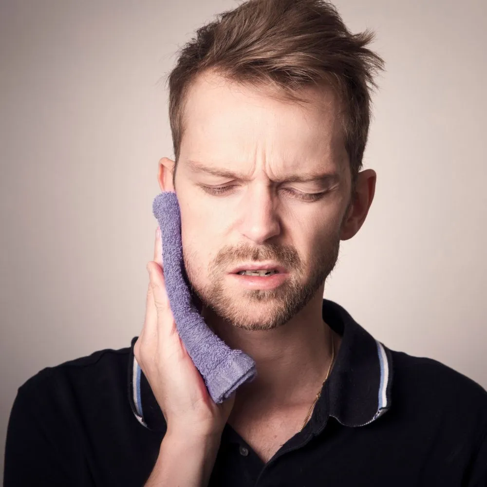 A young man pressing an ice pack to his cheek with eyes closed, showing visible discomfort from dental pain or swelling.