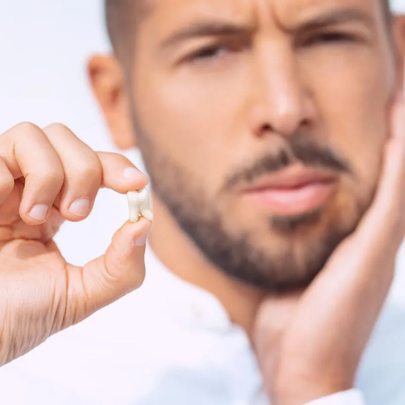 A man holding an extracted tooth in one hand while touching his cheek in discomfort, indicating recent dental pain or surgery.