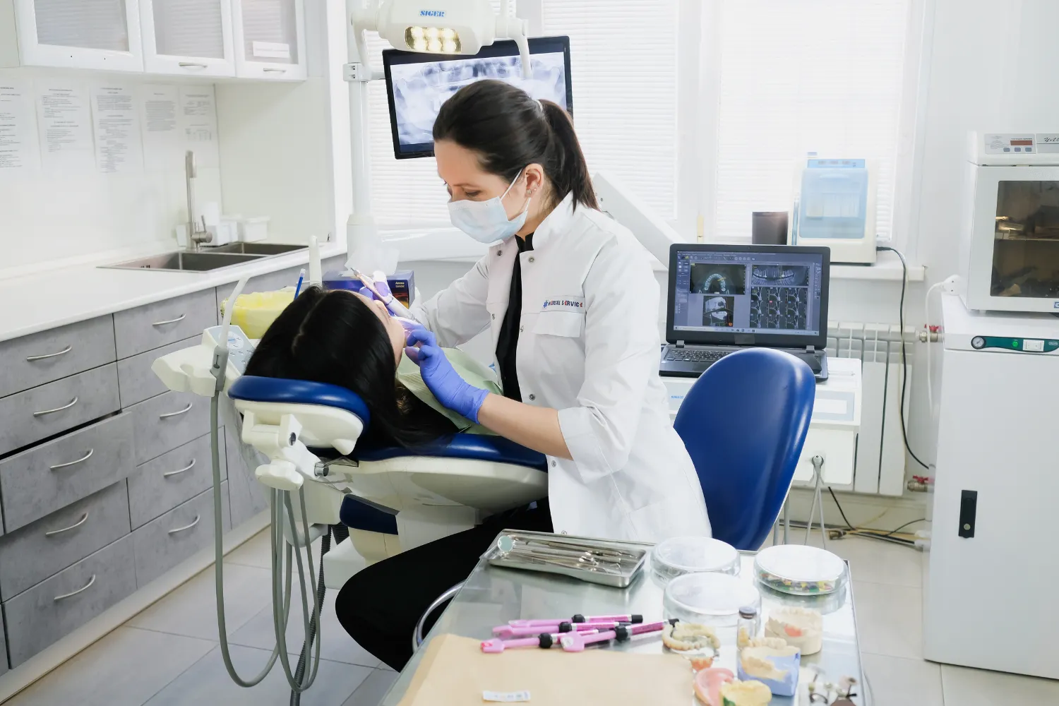 A female dentist in a white coat and mask performing a dental procedure on a patient in a modern operatory with X-rays on screen.