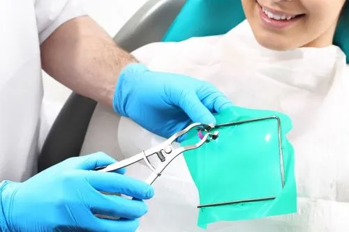 A dentist preparing a green rubber dam with forceps while a smiling patient sits in the dental chair, ready for isolated treatment.