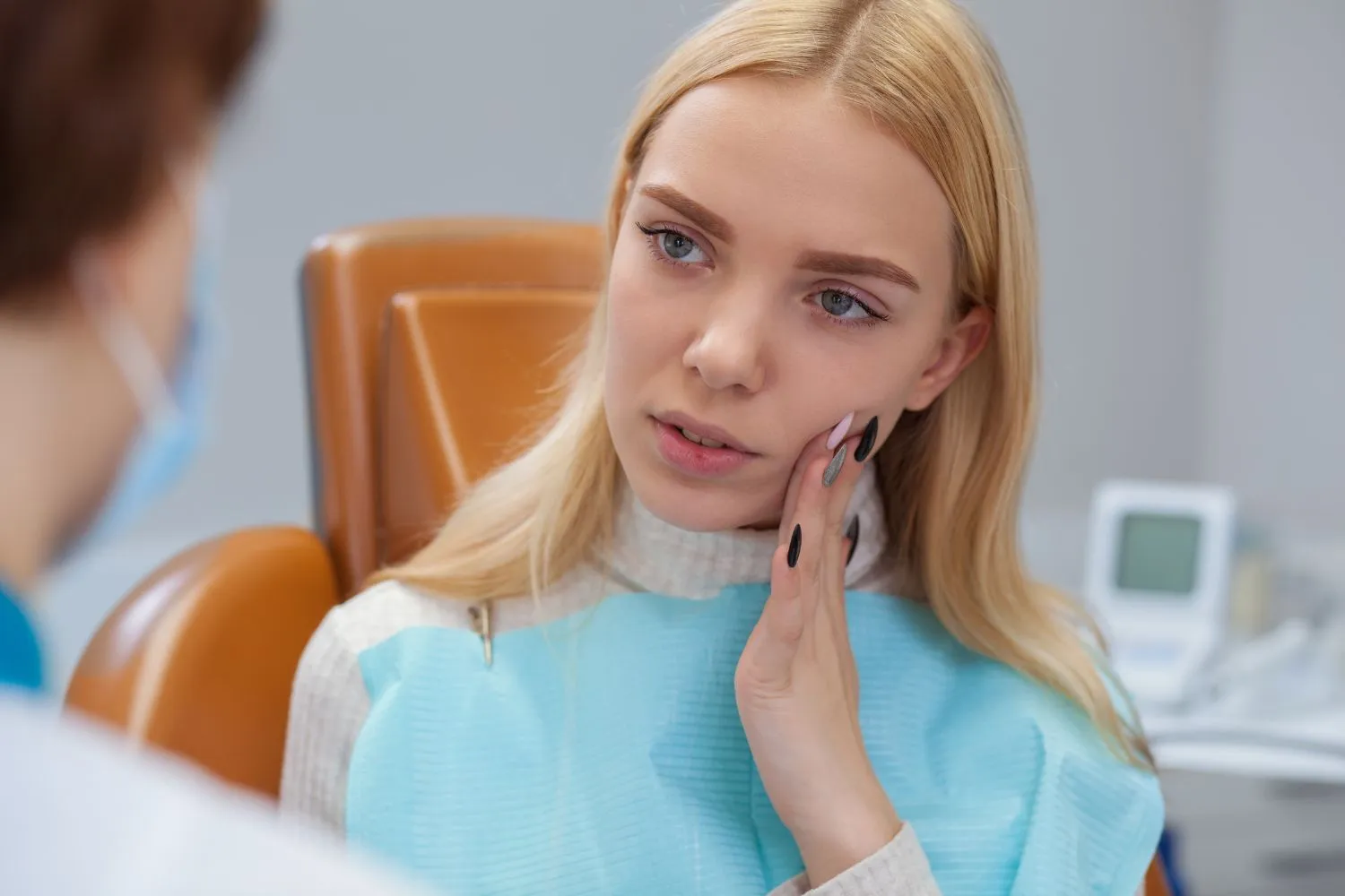 A young woman in a dental chair touching her cheek and talking to the dentist, expressing concern about tooth or jaw pain.