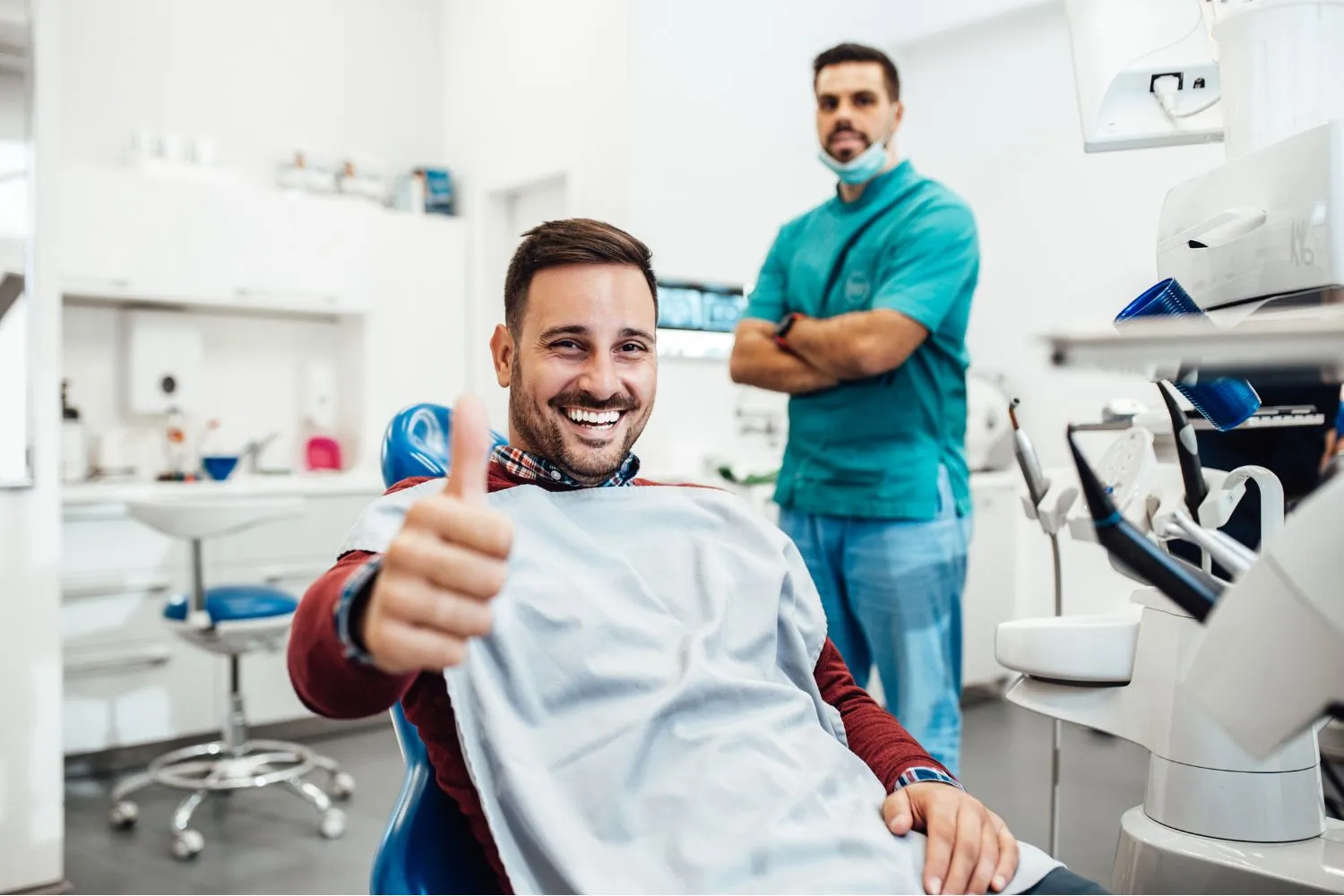 A smiling male patient giving a thumbs up while seated in a dental chair, with a confident dentist standing in the background.