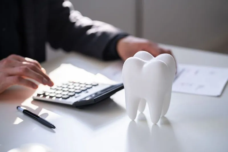 A person using a calculator at a desk with dental tooth models nearby, symbolizing financial planning or cost estimation for dental care.