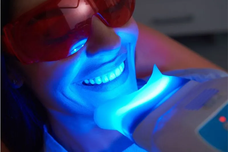 Close-up of a woman wearing red protective glasses undergoing a professional teeth whitening treatment using a blue LED light device.