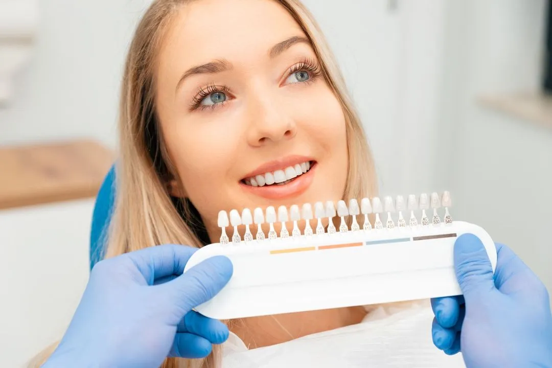 A happy blonde woman in the dental chair looking at a shade guide held by a dentist for tooth color matching.