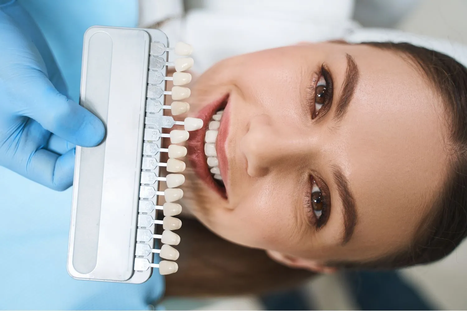 A patient in a blue bib smiling as a dentist compares her teeth to a full spectrum shade guide for veneer or whitening treatment.