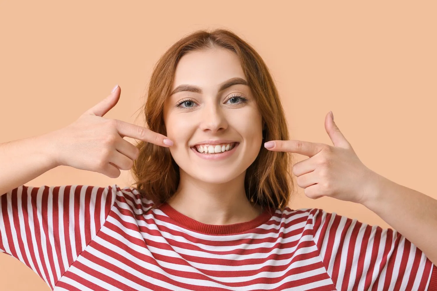 A cheerful young woman in a striped shirt pointing at her white teeth, confidently showing off her bright smile.