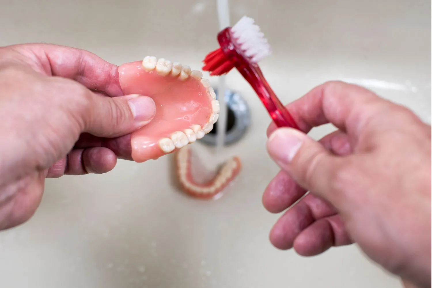 Hands holding a red denture brush and upper denture over a sink, demonstrating proper hygiene for removable dental appliances.