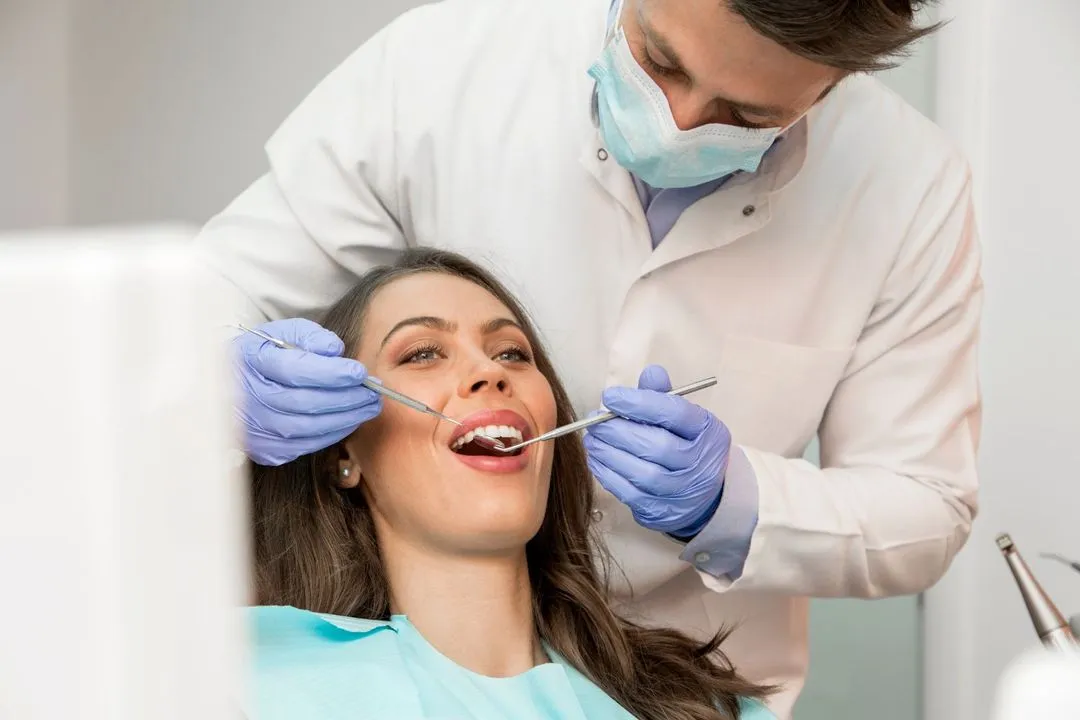 A female patient receiving a standard dental exam as a masked dentist uses instruments to inspect her teeth.