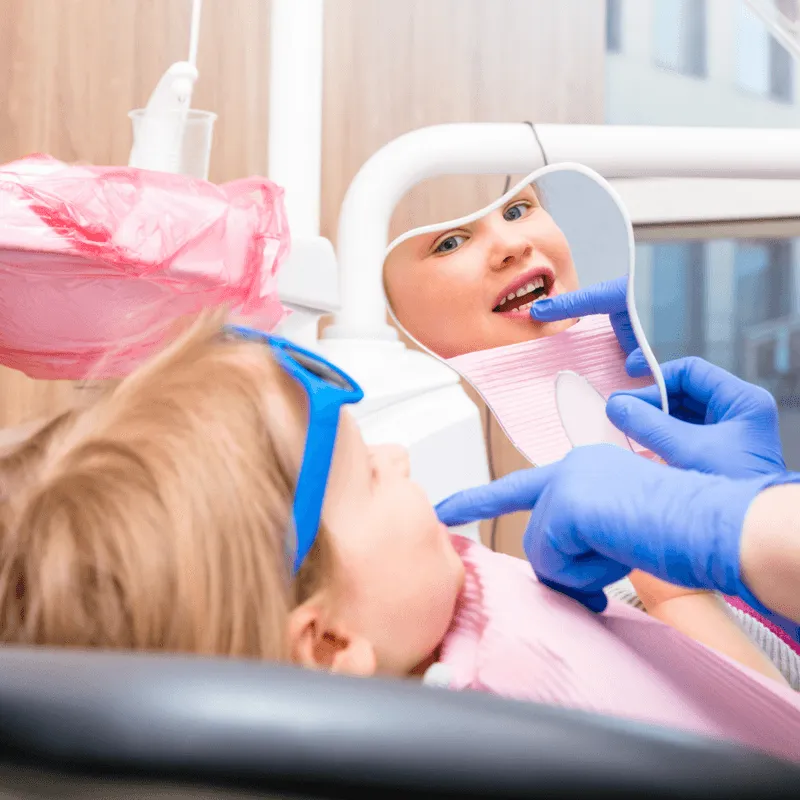 A young girl wearing blue glasses pointing at her reflection in a dental mirror during her appointment.