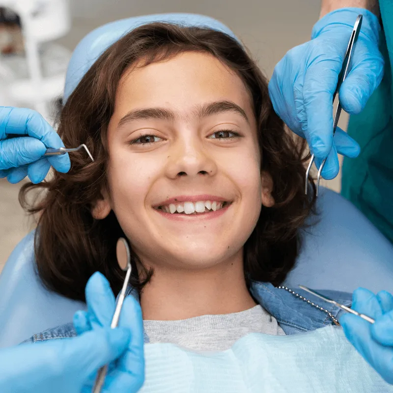 A smiling girl receiving a dental checkup with multiple gloved hands holding dental instruments around her.