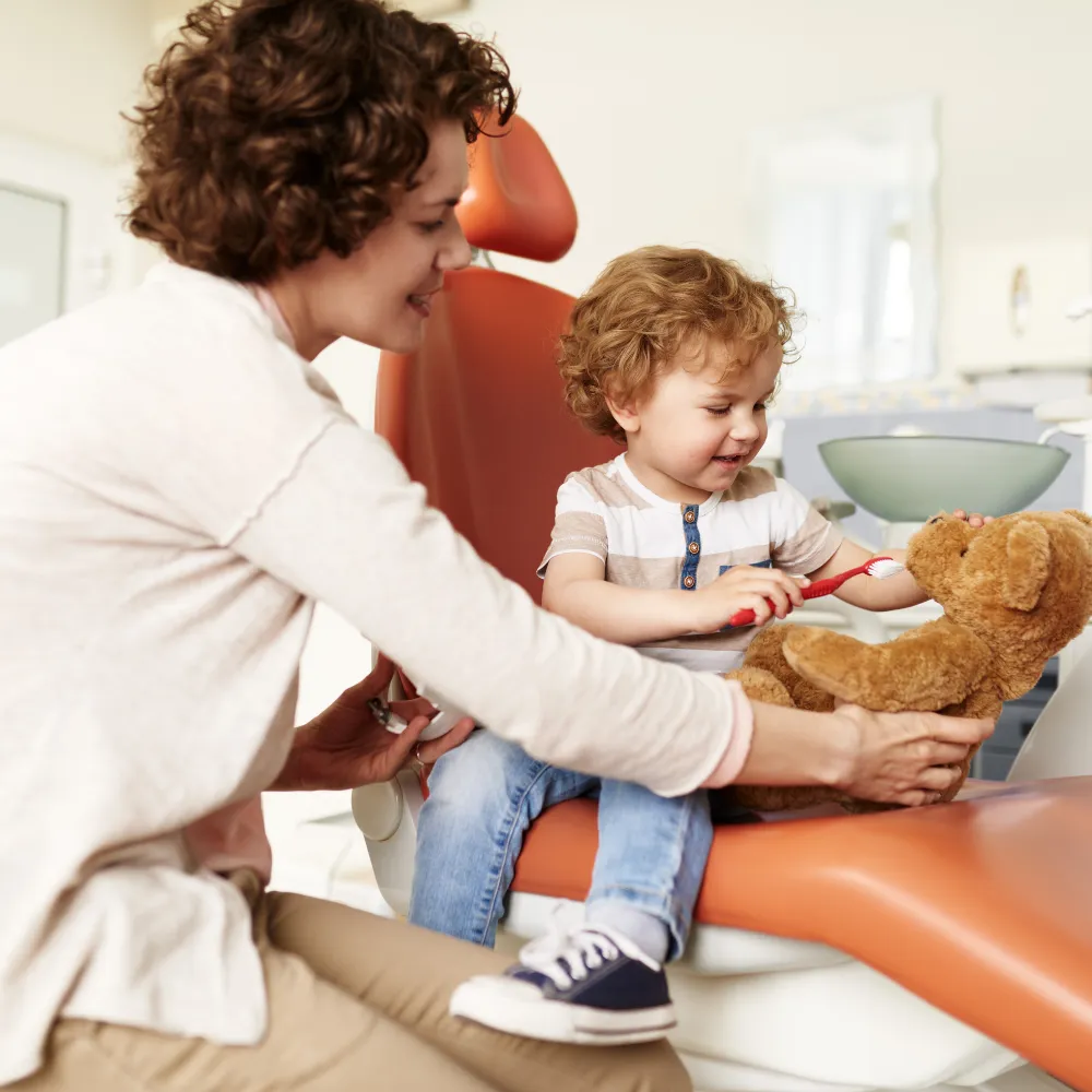 A young child in a dental chair brushing a teddy bear's teeth with help from a female dentist, highlighting child-friendly dental care.
