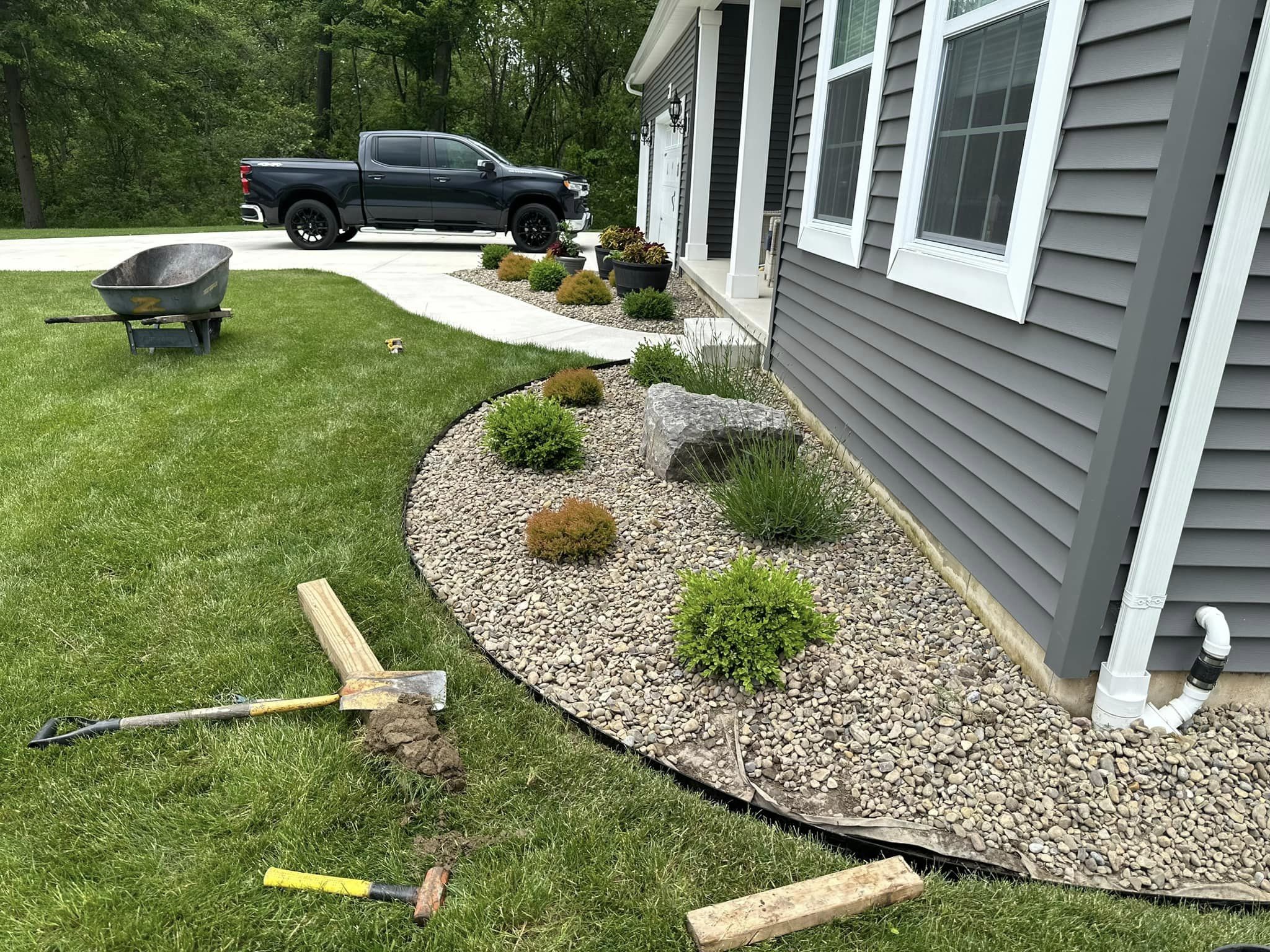 A front yard garden bed with newly installed steel edging separating a rock-filled bed with small shrubs from the grass lawn. A wheelbarrow and tools are in the foreground.