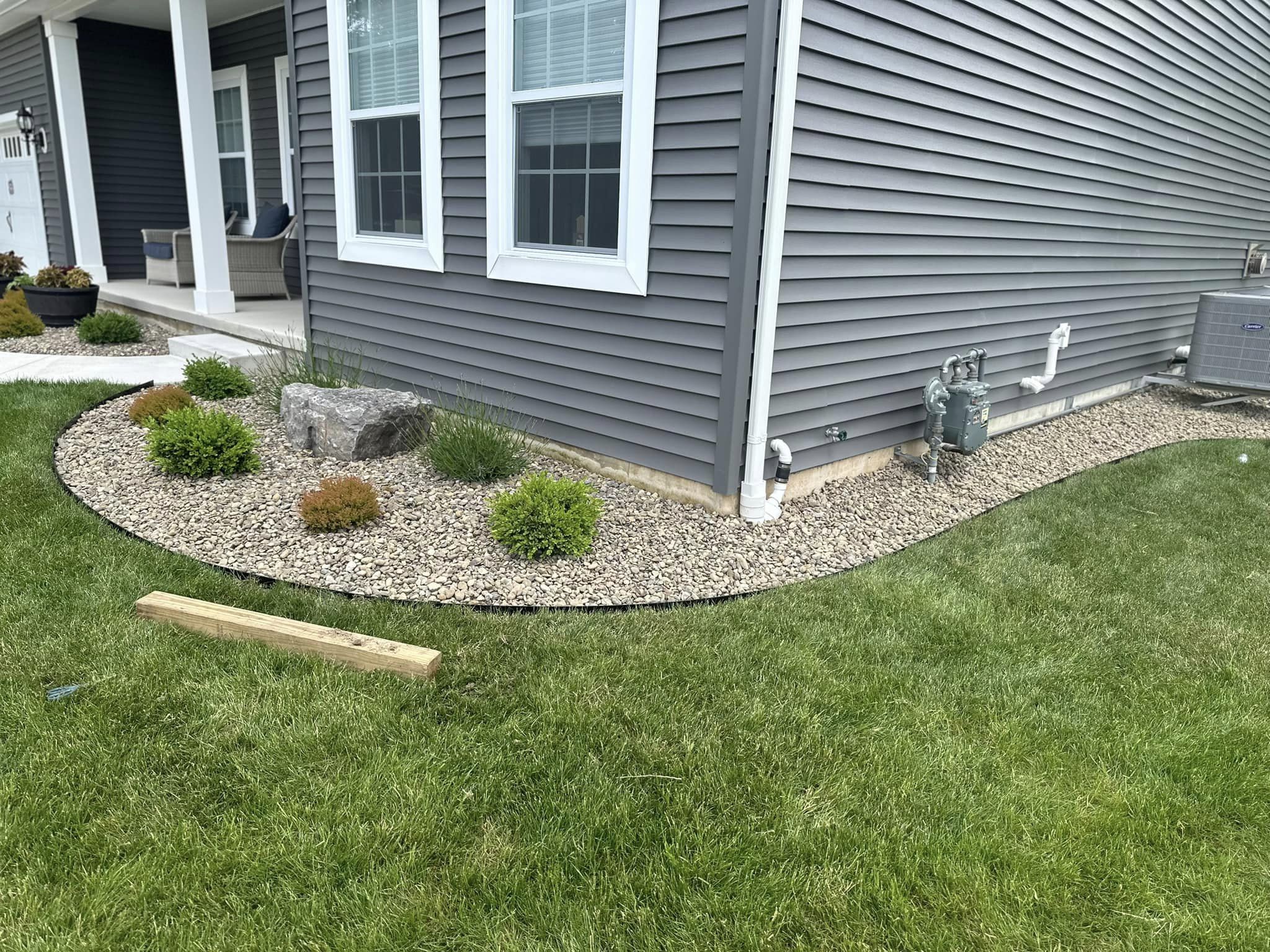 A front yard garden with freshly installed steel edging creating a clean line between the rock-filled bed with shrubs and the grass lawn. The house's exterior is visible in the background.