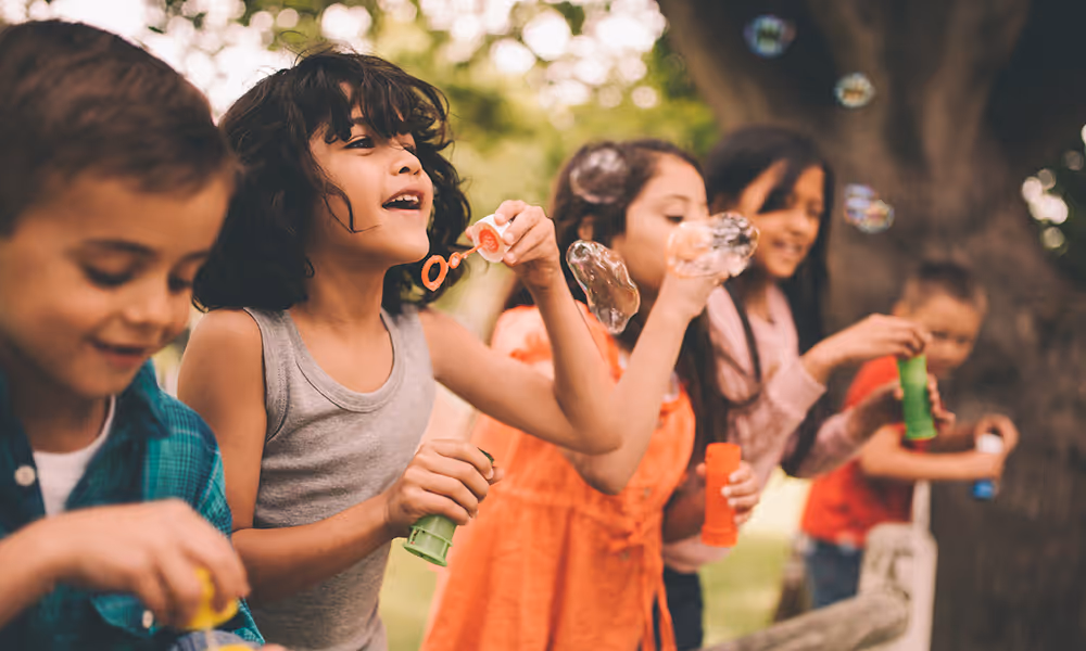 A group of diverse children are outdoors in a grassy area, smiling and laughing as they blow soap bubbles.