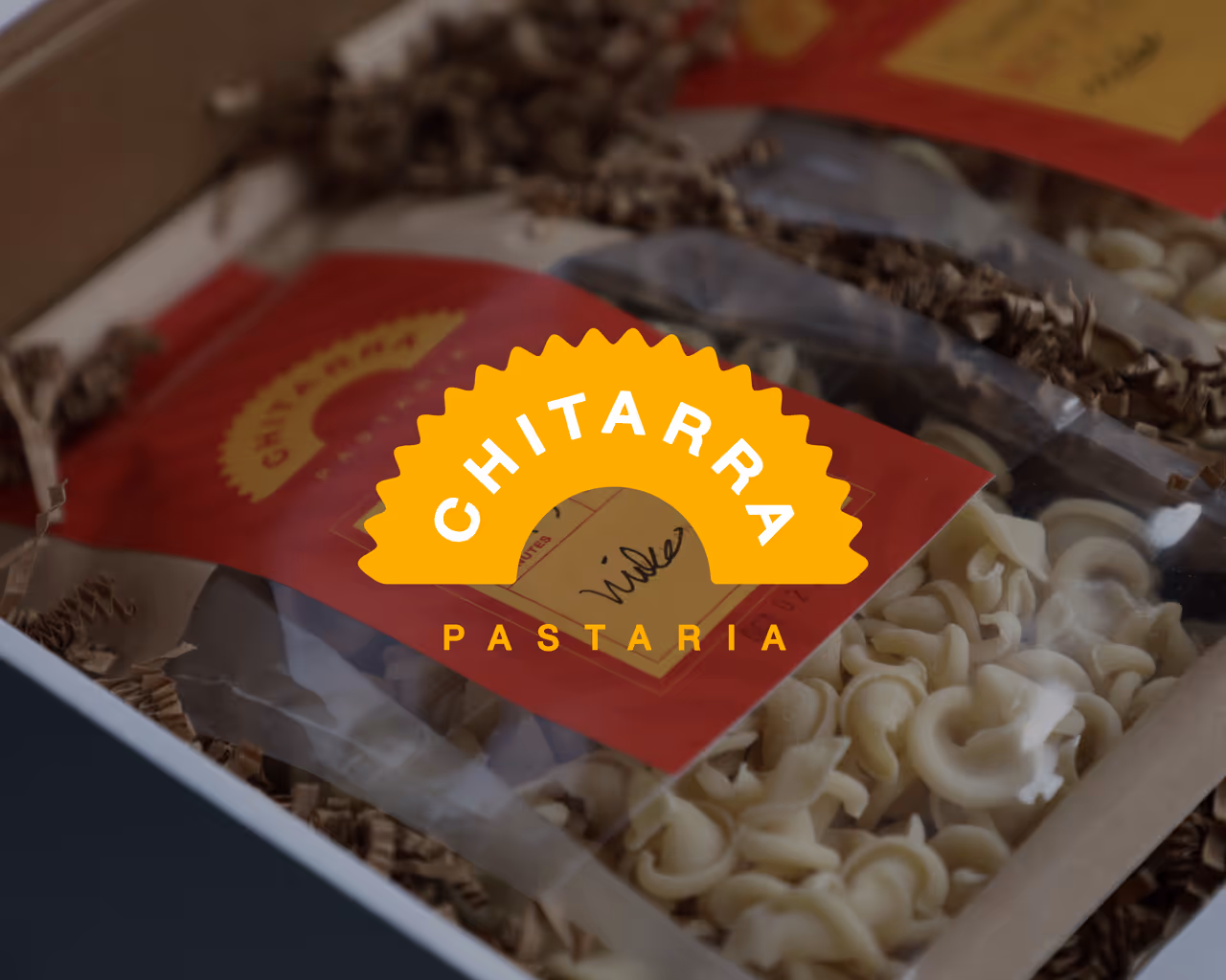Chitarra Pastaria logo in orange overlaid on a photo of packaged artisanal pasta in clear bags with red labels inside a gift box.