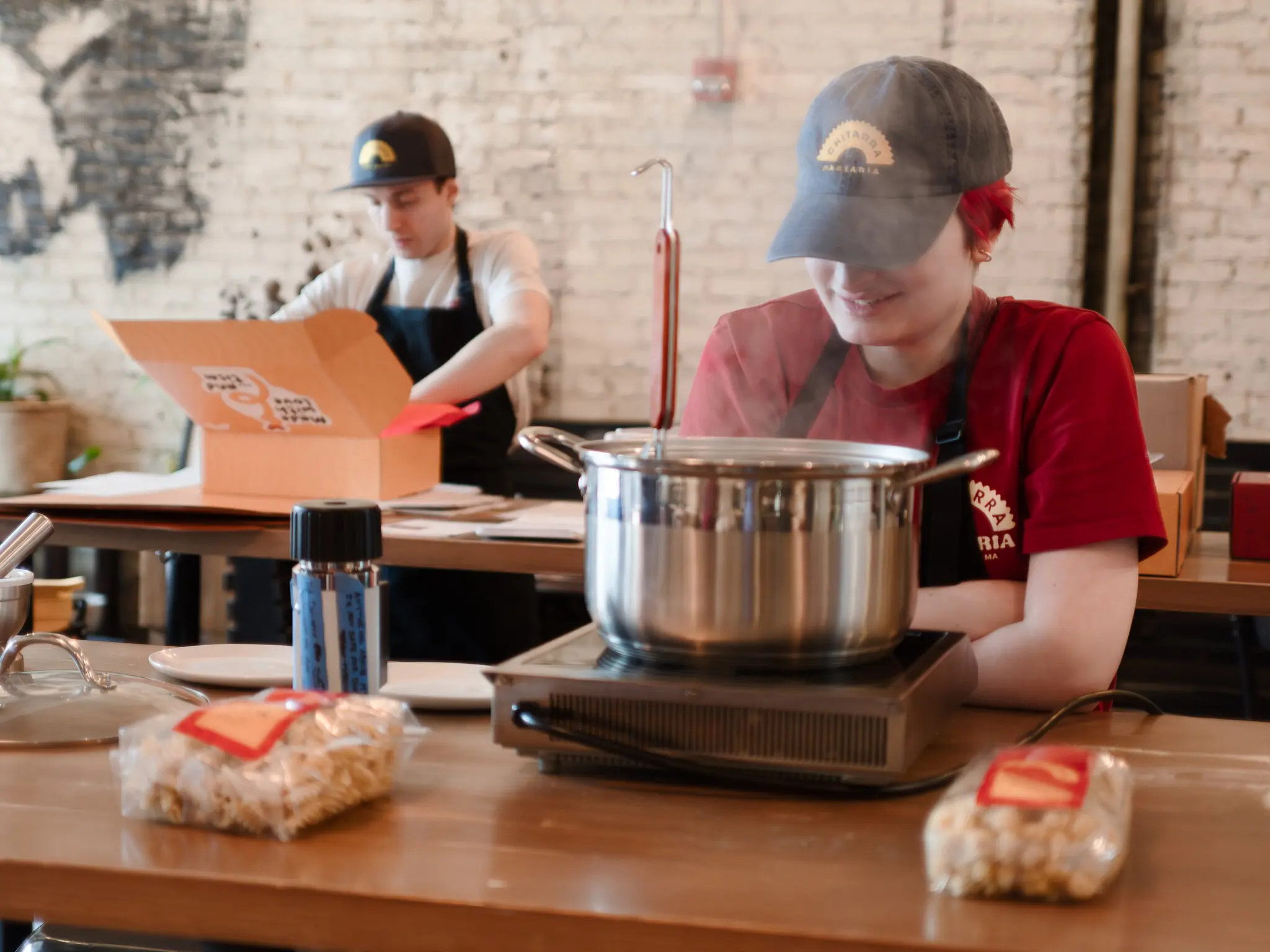 A worker in a red T-shirt supervises a boiling pot while another, in a white T-shirt and blue apron, packs a cardboard box.