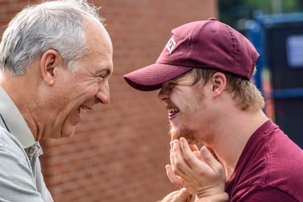 A candid, heartwarming photo of an older man and a younger man with Down syndrome laughing together outdoors. They are face-to-face in a close, affectionate moment.