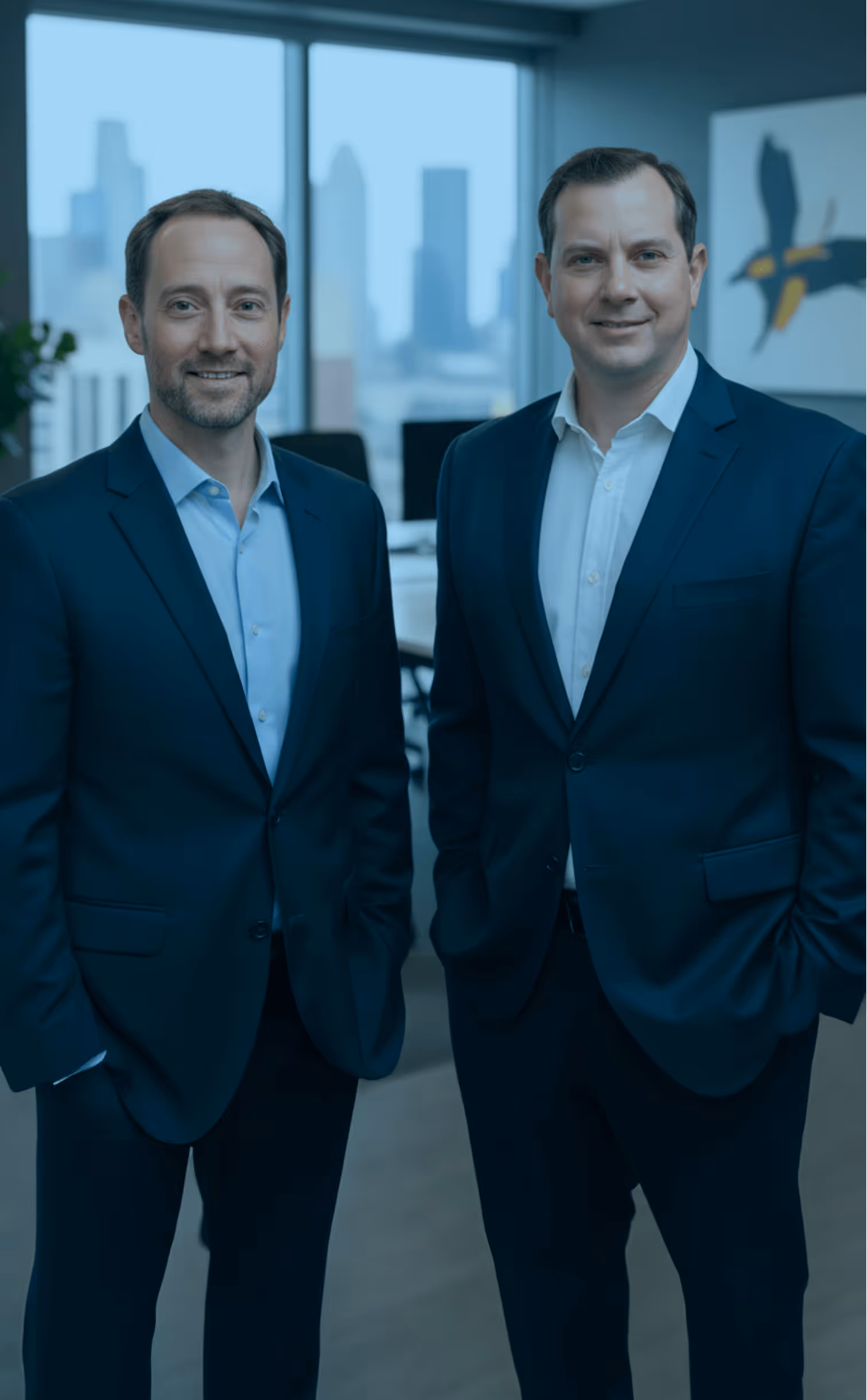 Two men in business suits smiling in a modern office with large windows and city skyline in the background.