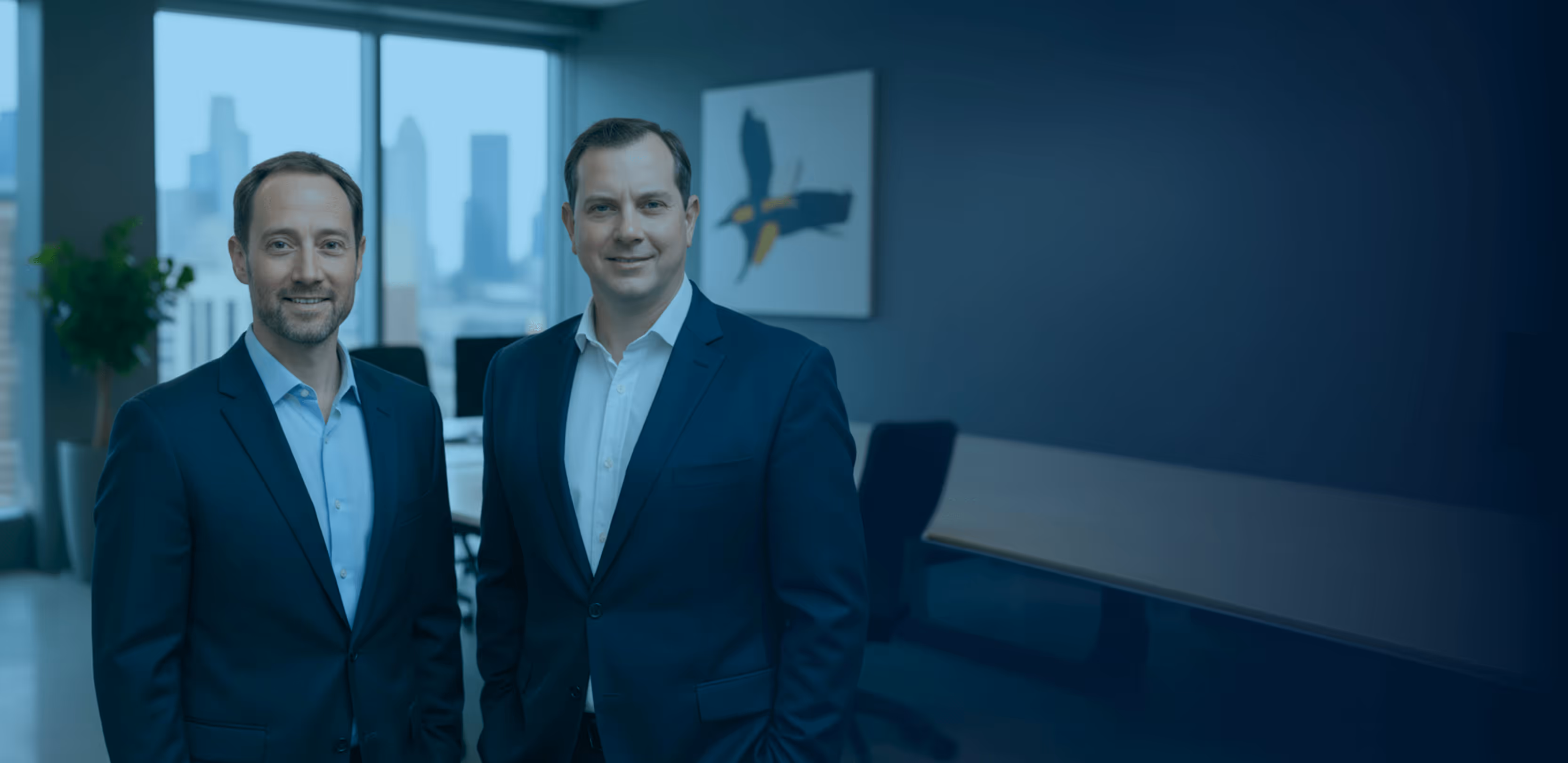 Two men in suits standing in a modern office with a city skyline visible through large windows.