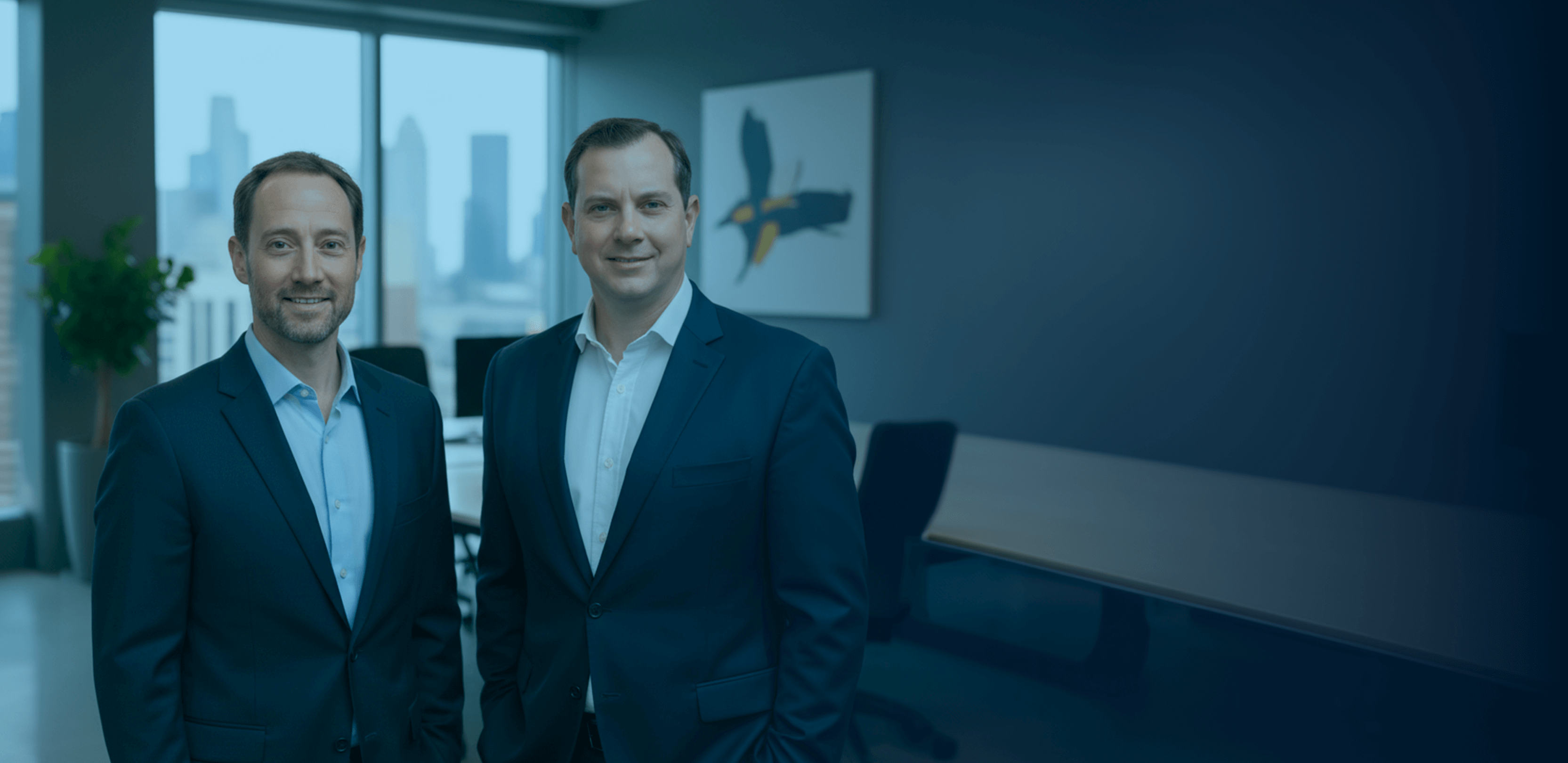 Two men in suits standing in a modern office with a city skyline visible through large windows.