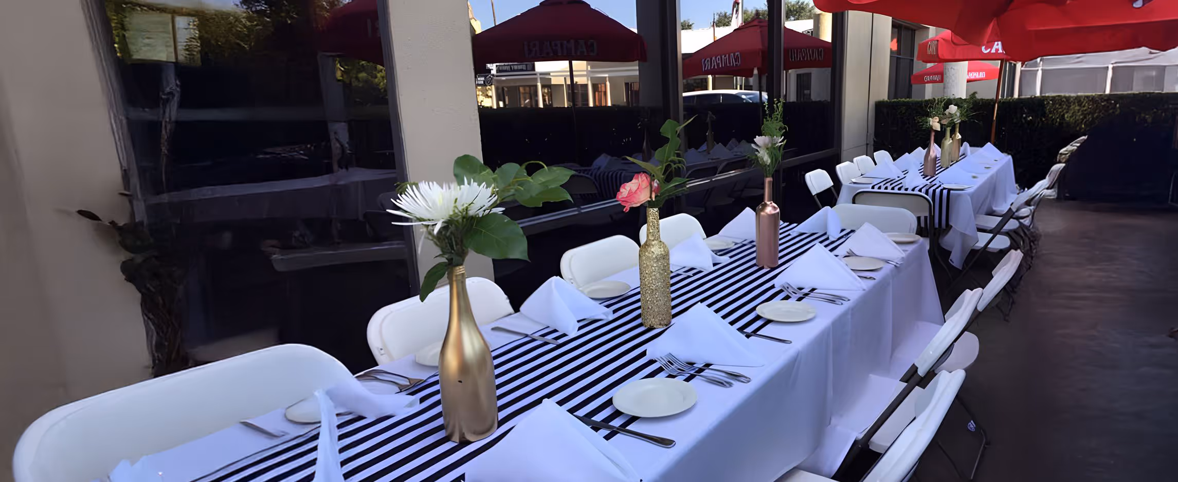 Elegant outdoor table setting with white chairs and striped tablecloths. Gold vases hold flowers, under vibrant red umbrellas.