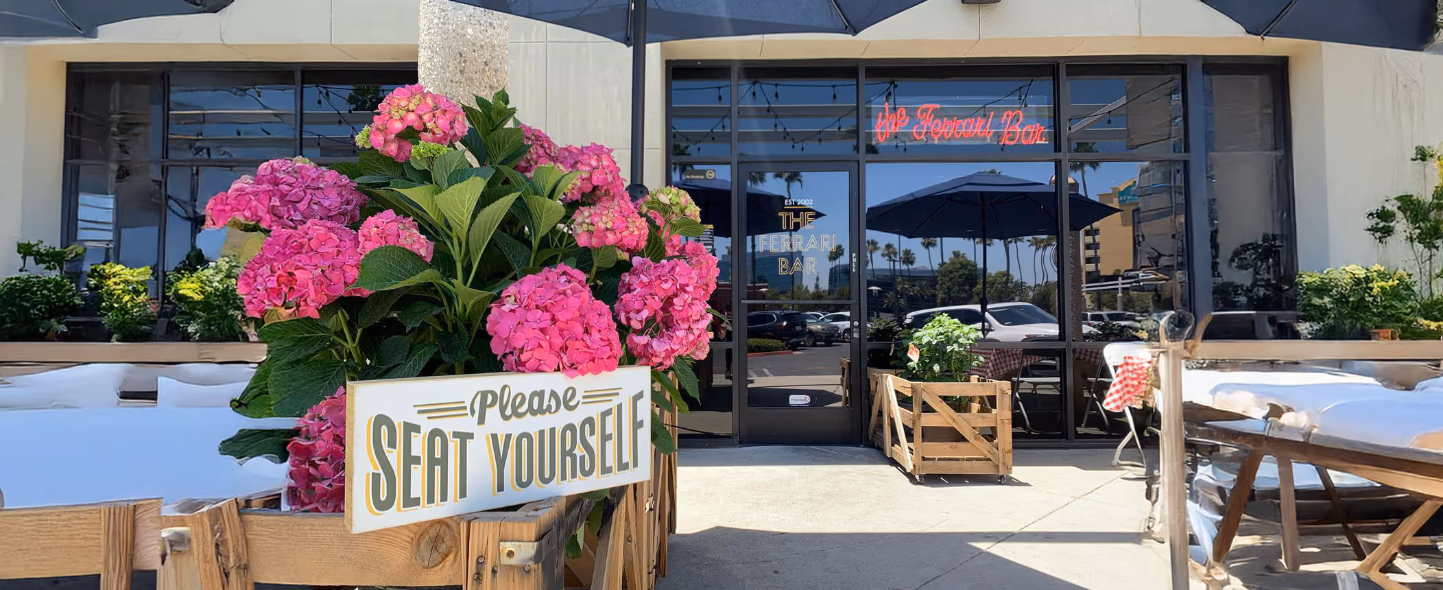 Outdoor view of a restaurant's entrance with pink flowers in the foreground and a sign reading "Please Seat Yourself." Tables and chairs are set under a sunny sky, creating a welcoming atmosphere.