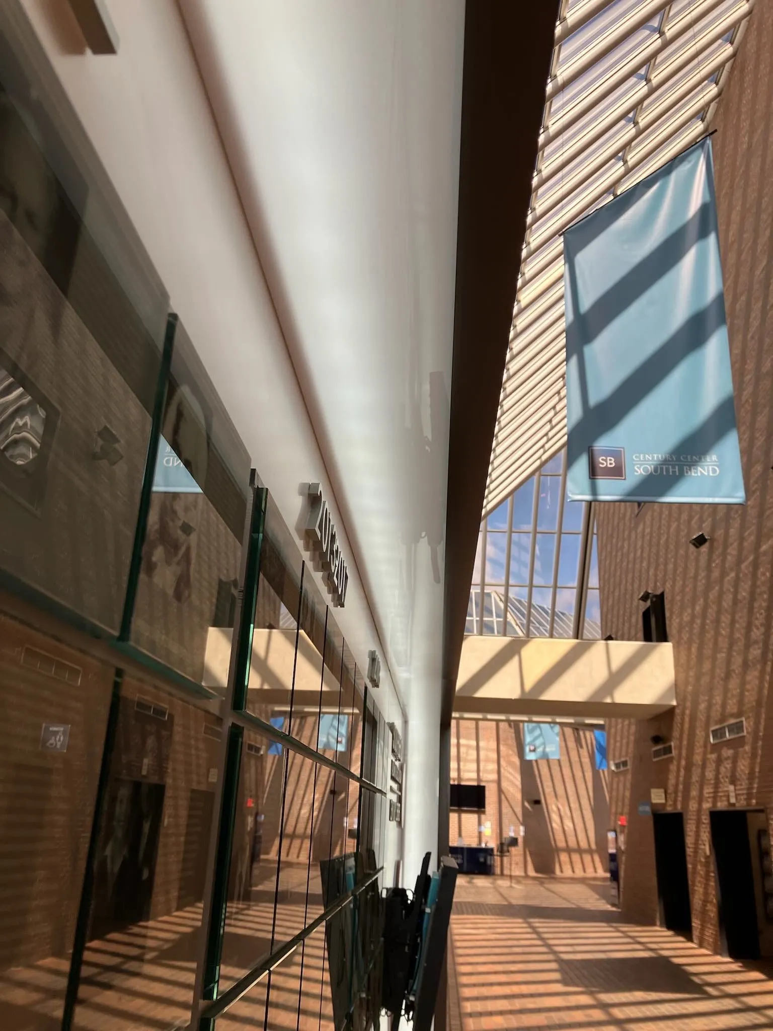 Interior view of a modern building lobby with large windows casting striped shadows on brick walls and floor, and a blue banner reading 'Century Center South Bend'.