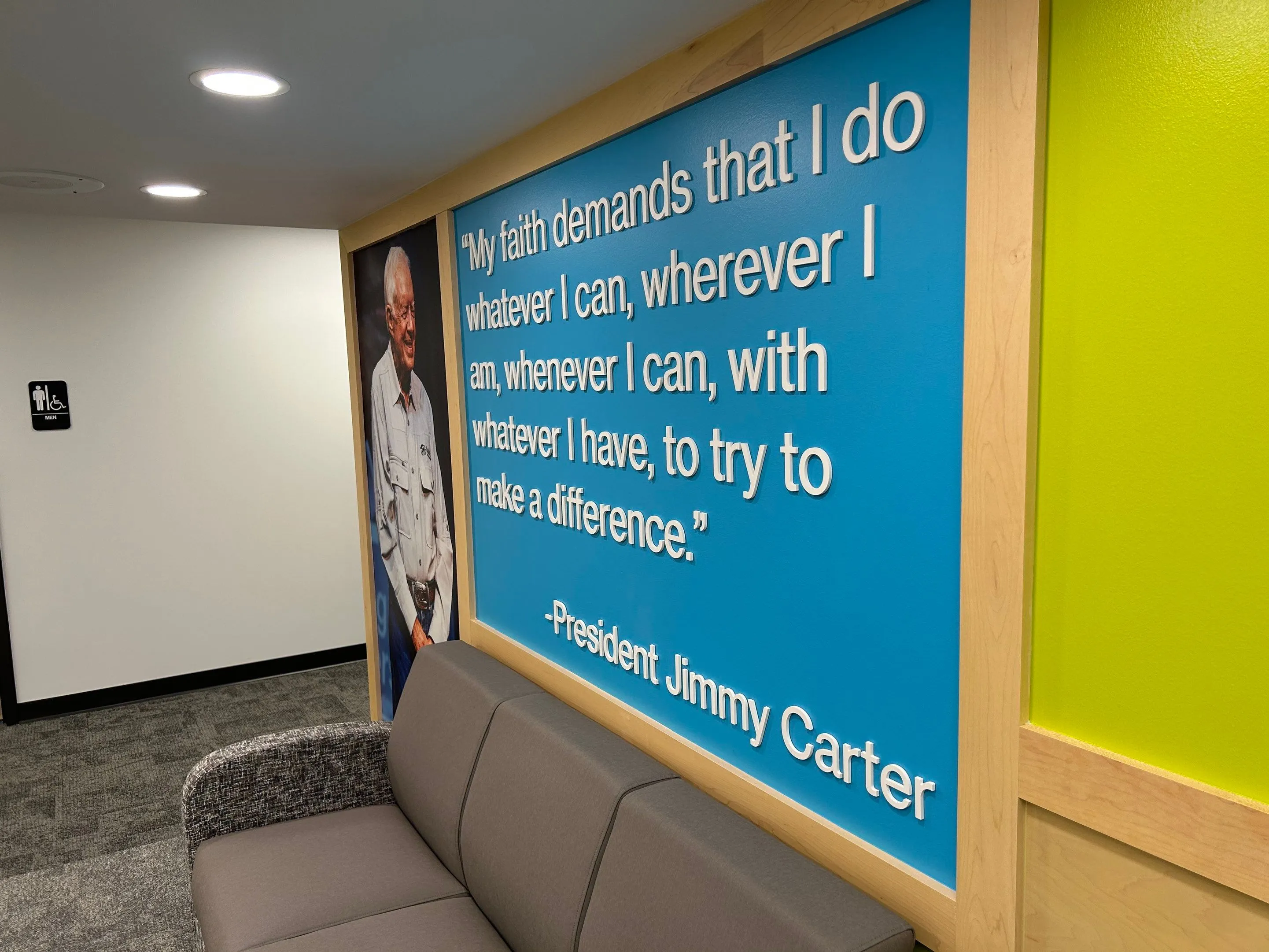 Wall with a blue framed quote by President Jimmy Carter next to a photo of him and a gray couch below; quote reads: 'My faith demands that I do whatever I can, wherever I am, whenever I can, with whatever I have, to try to make a difference.'
