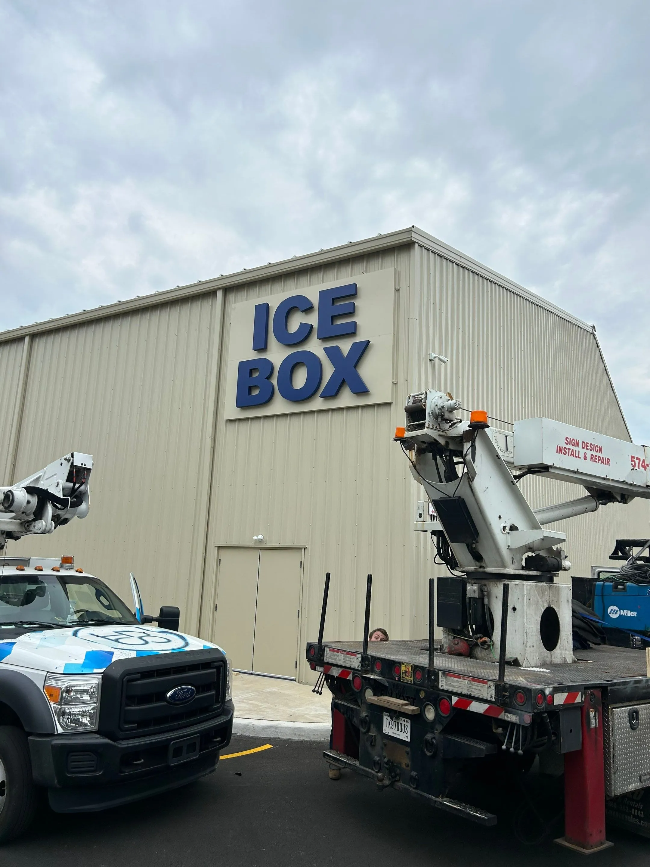 Two trucks with equipment parked outside a beige industrial building with large blue letters reading 'ICE BOX' on the wall.