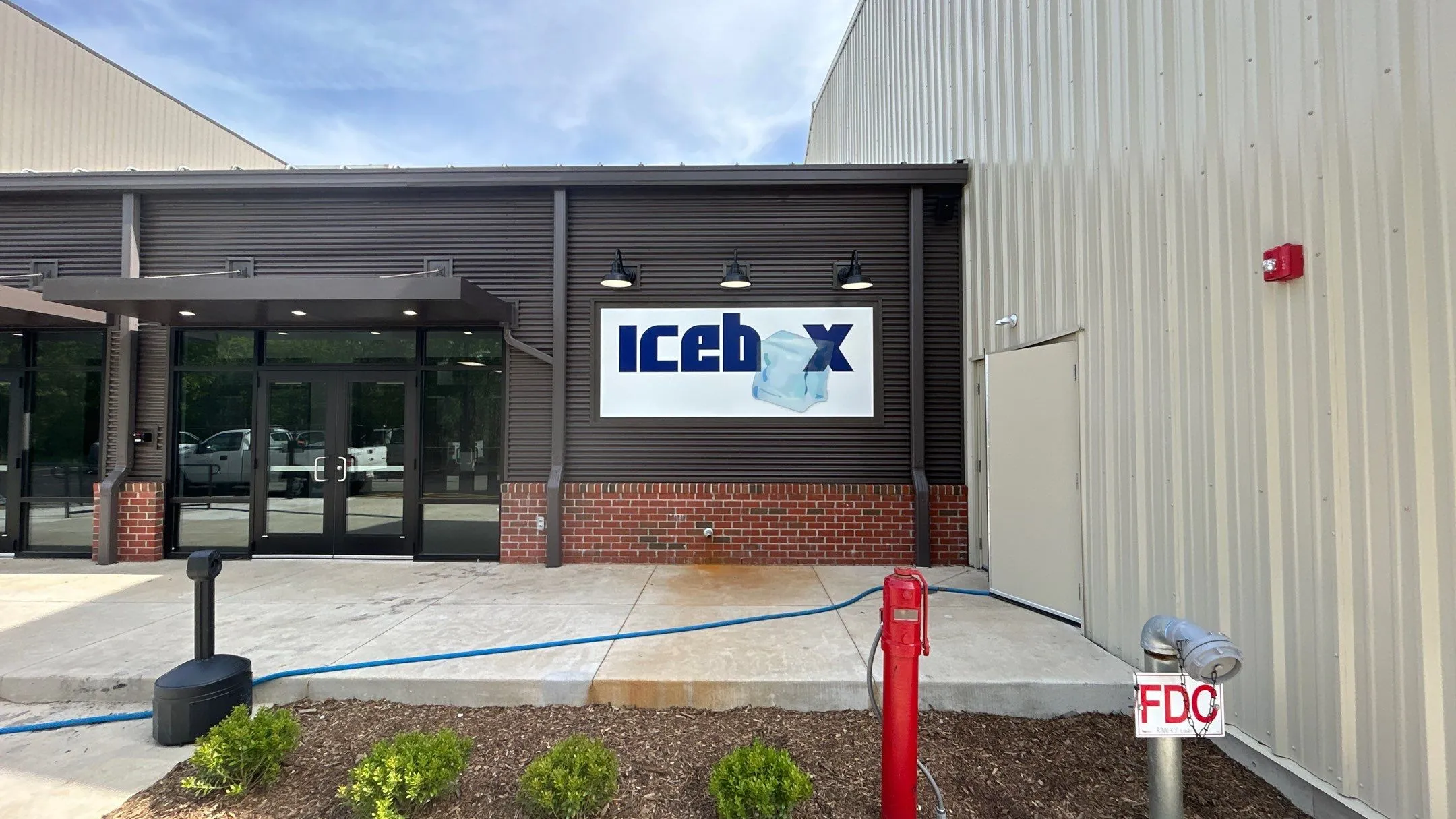 Exterior of a commercial building with dark brown siding, brick base, glass double doors, and a sign with the text 'iceb x' and an ice cube graphic.