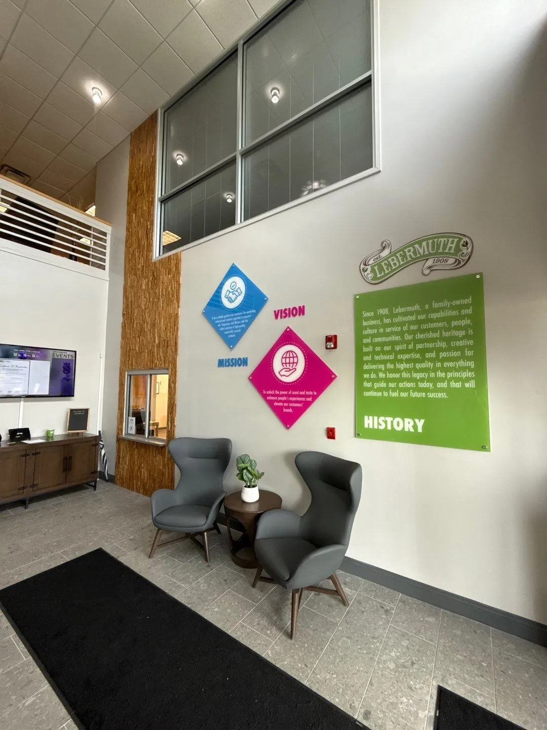 Modern office lobby with two gray lounge chairs and a small round table with a plant, wall displays showing company mission, vision, and history in colored panels.