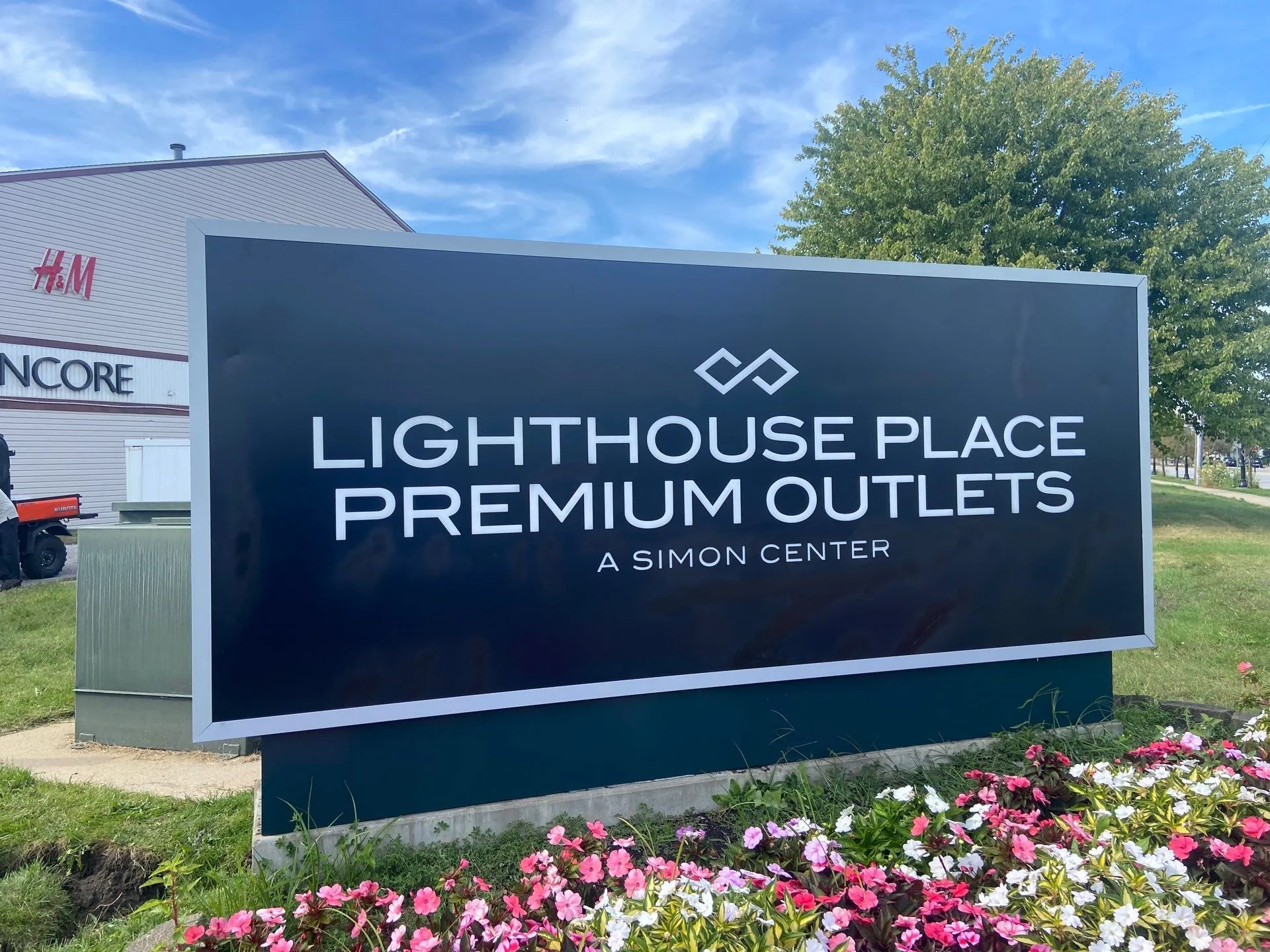 Black sign for Lighthouse Place Premium Outlets at a Simon Center surrounded by grass and pink, white, and red flowers.