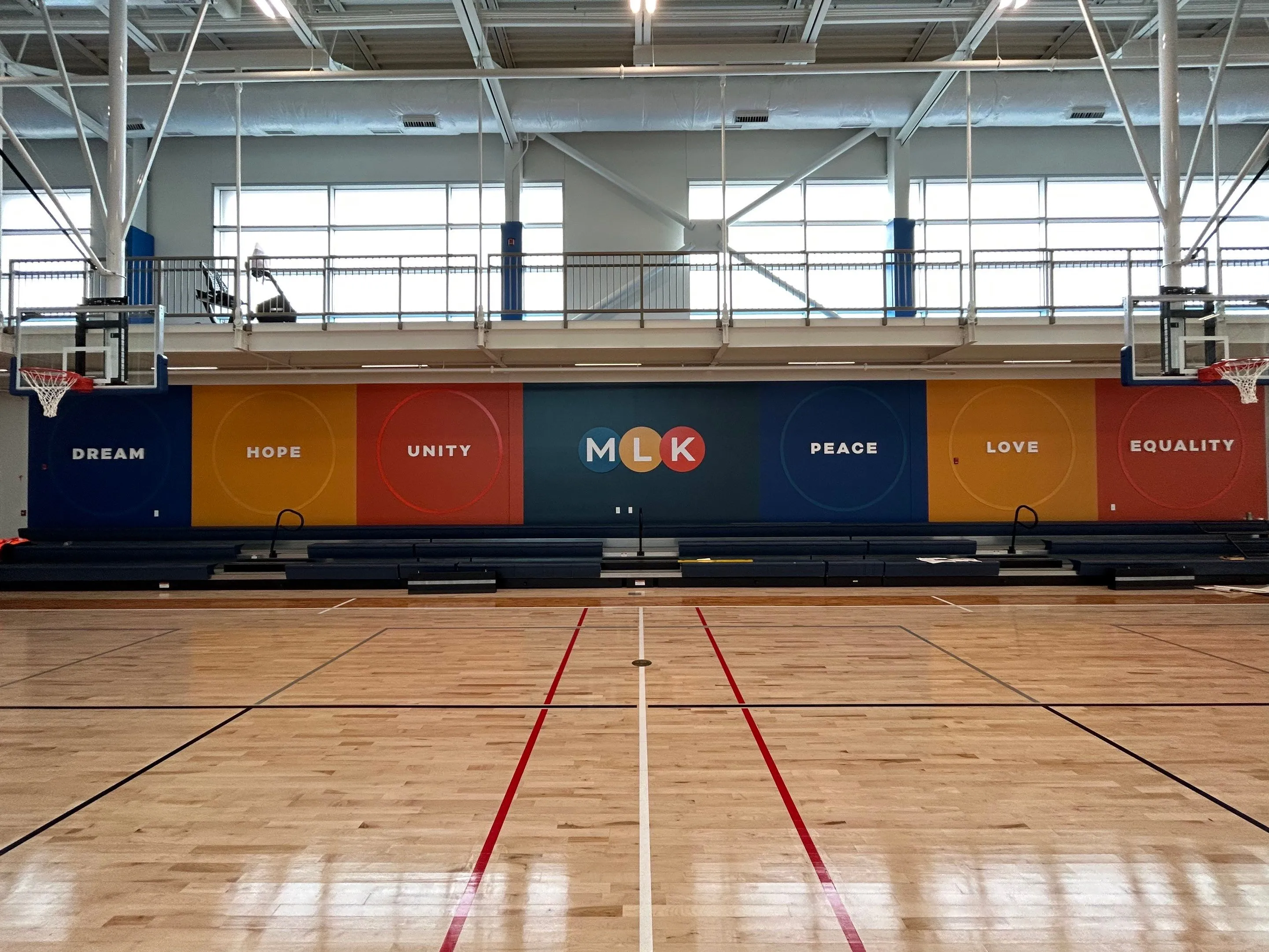 Indoor basketball court with a mural on the far wall displaying the words DREAM, HOPE, UNITY, MLK, PEACE, LOVE, and EQUALITY in bold letters.