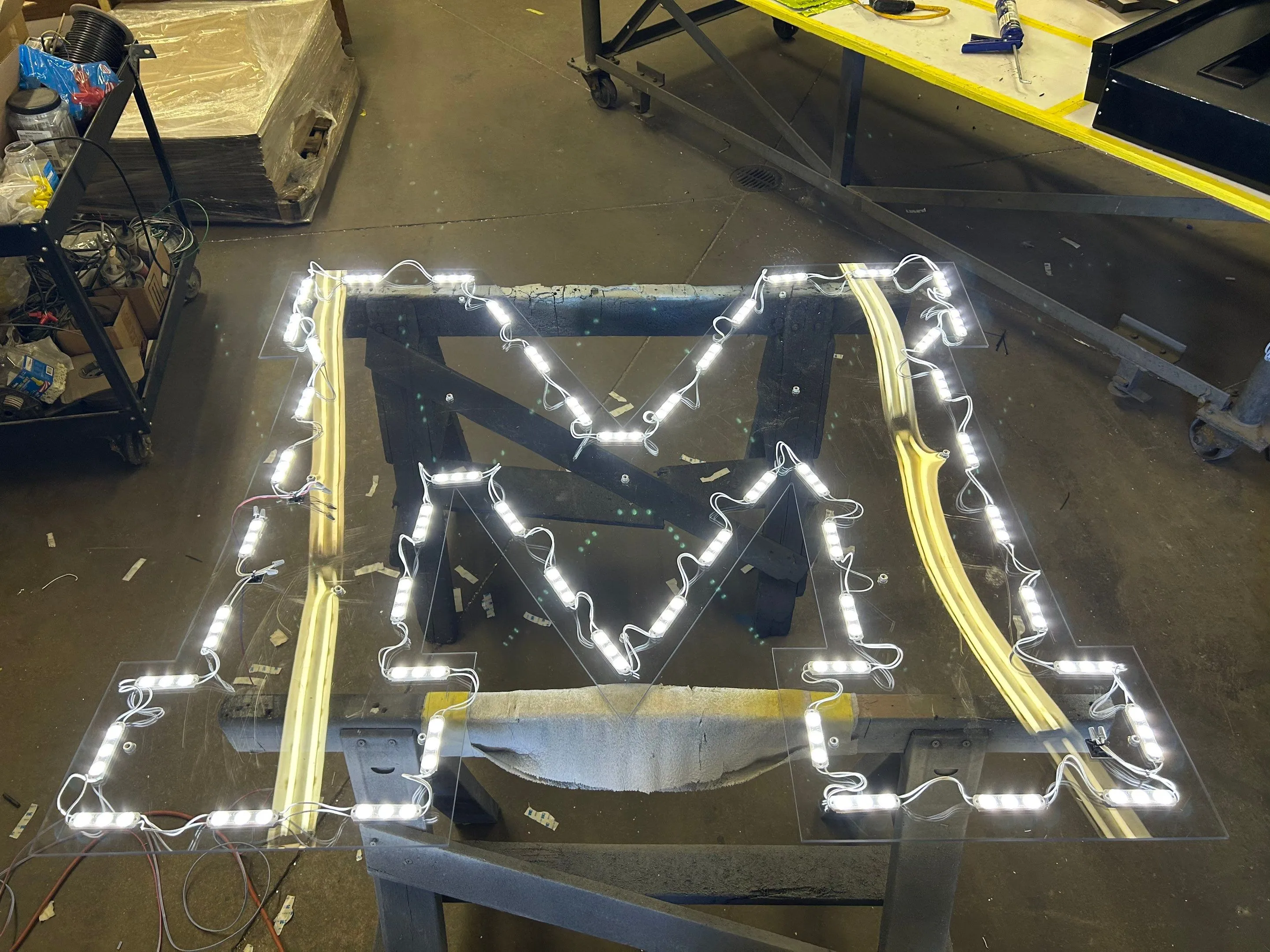 Large illuminated letter M with LED lights mounted on a transparent panel resting on a stand in a workshop.
