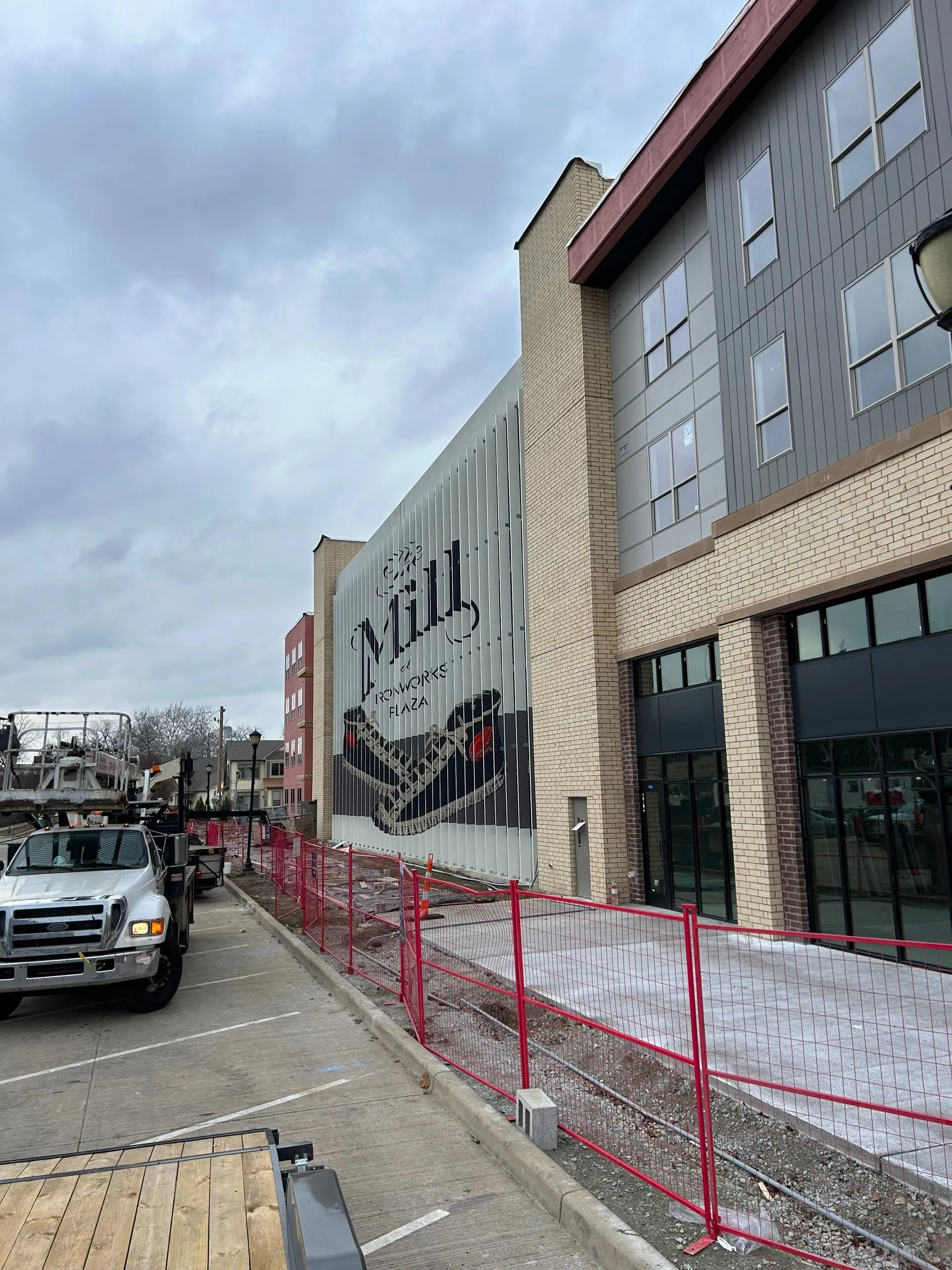 Street view of a building under construction with a large mural reading 'The Mill at Ironworks Plaza' featuring a black sneaker illustration, with construction fences and trucks nearby.