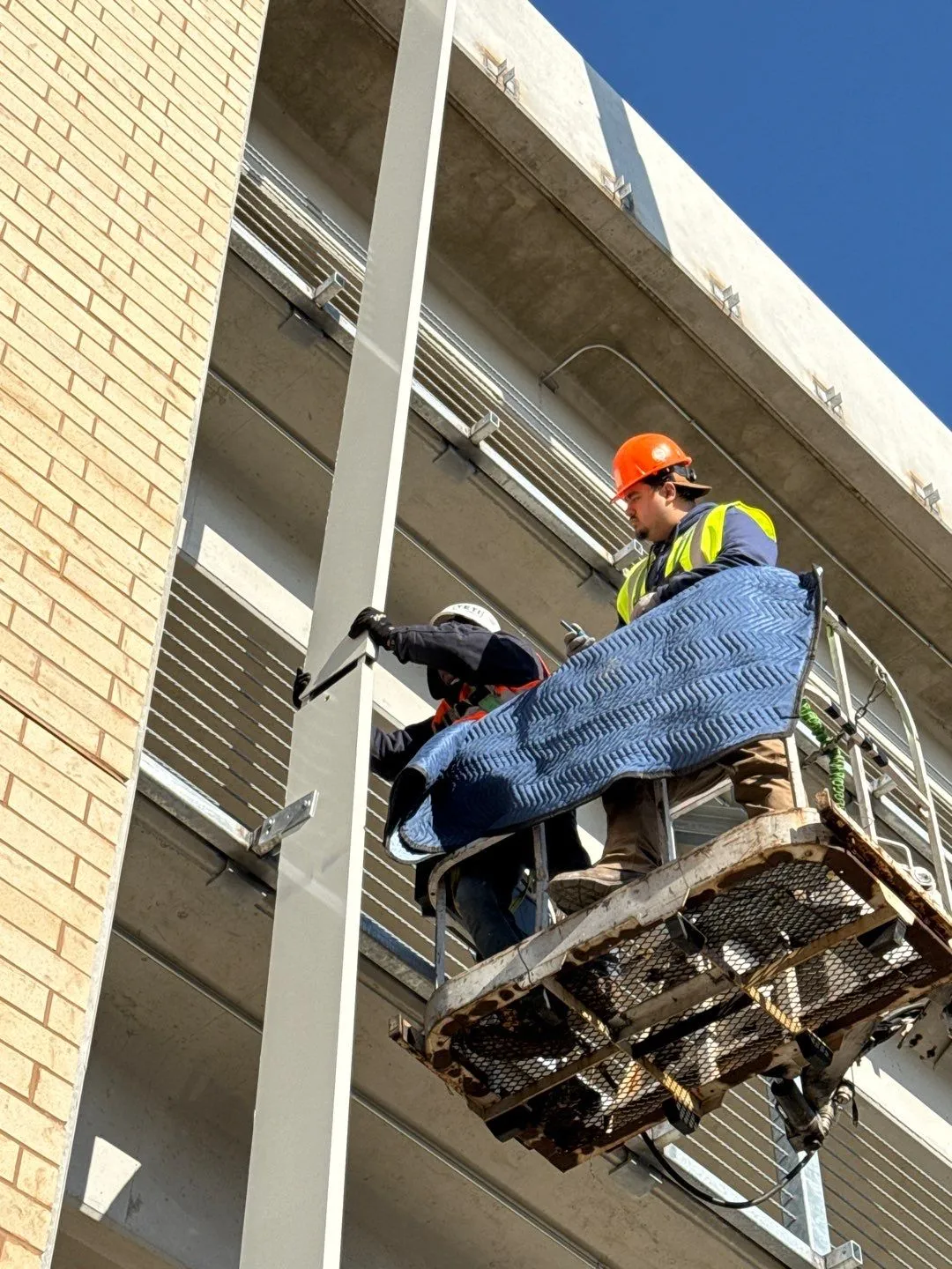 Two construction workers wearing helmets and safety vests working on a suspended platform on a building exterior against a clear blue sky.