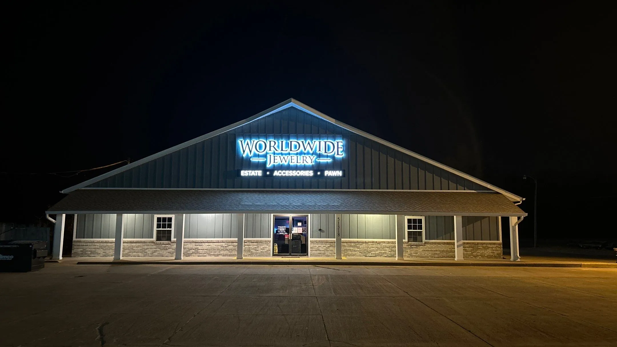 Lit storefront of Worldwide Jewelry with signs for estate, accessories, and pawn services at night.