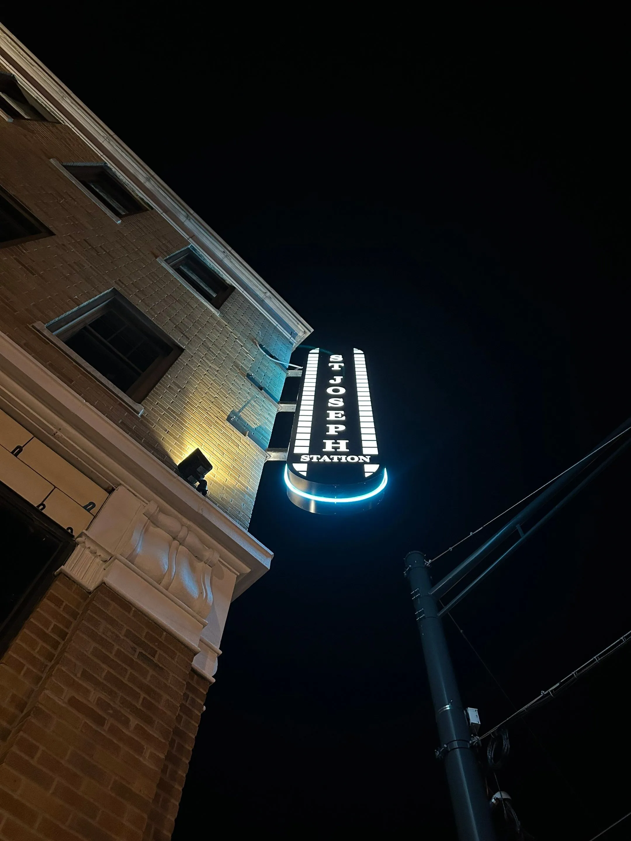 Illuminated vertical sign reading 'ST JOSEPH STATION' attached to a brick building at night.