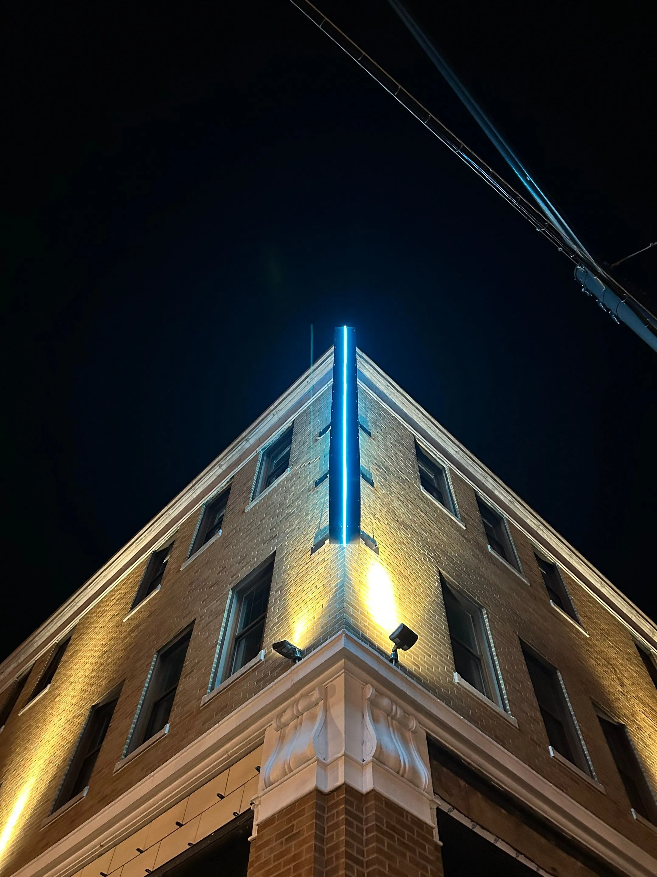 Upward view of a brick building corner with illuminated blue vertical light strip and yellow exterior lights at night.