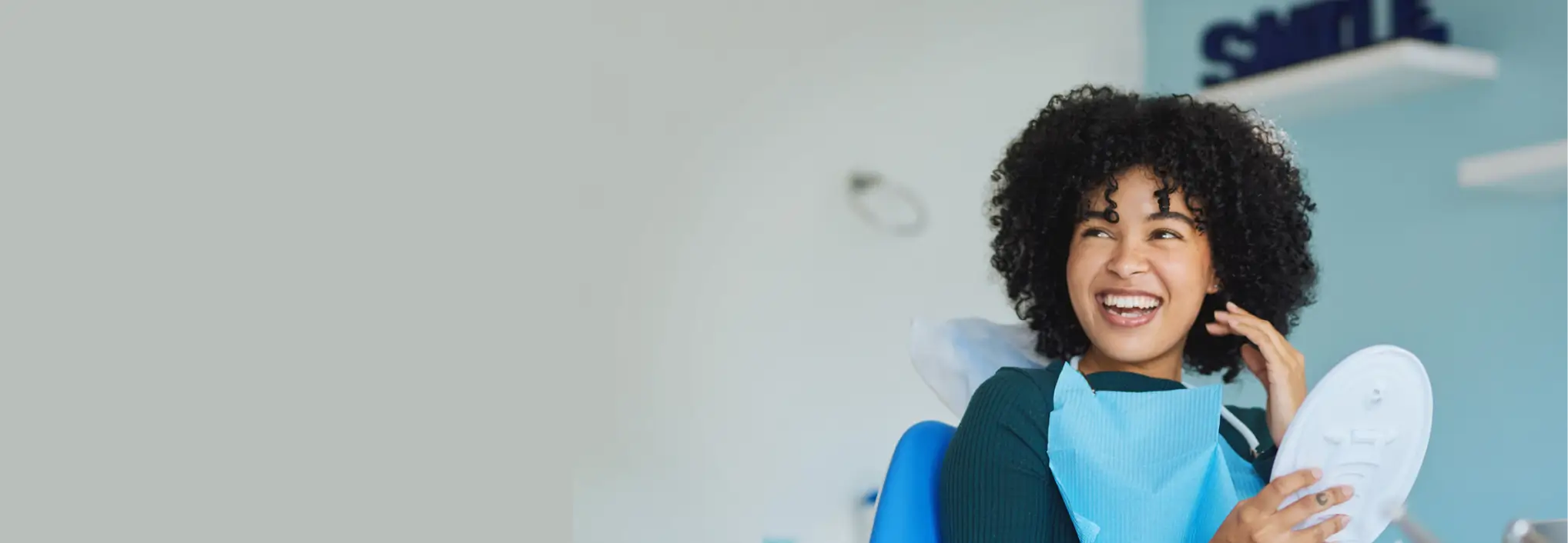 Smiling woman with curly hair sitting in a dental chair holding a mirror and wearing a dental bib.
