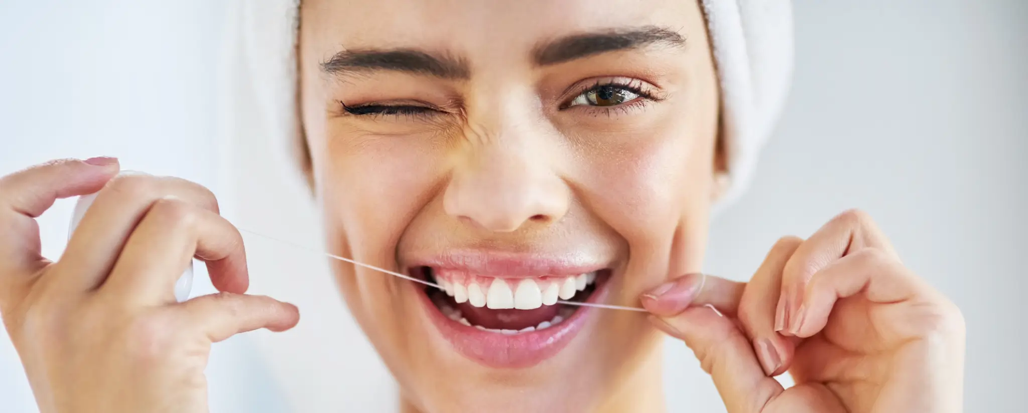 Close-up of a smiling woman with a towel wrapped around her head, flossing her teeth and winking.