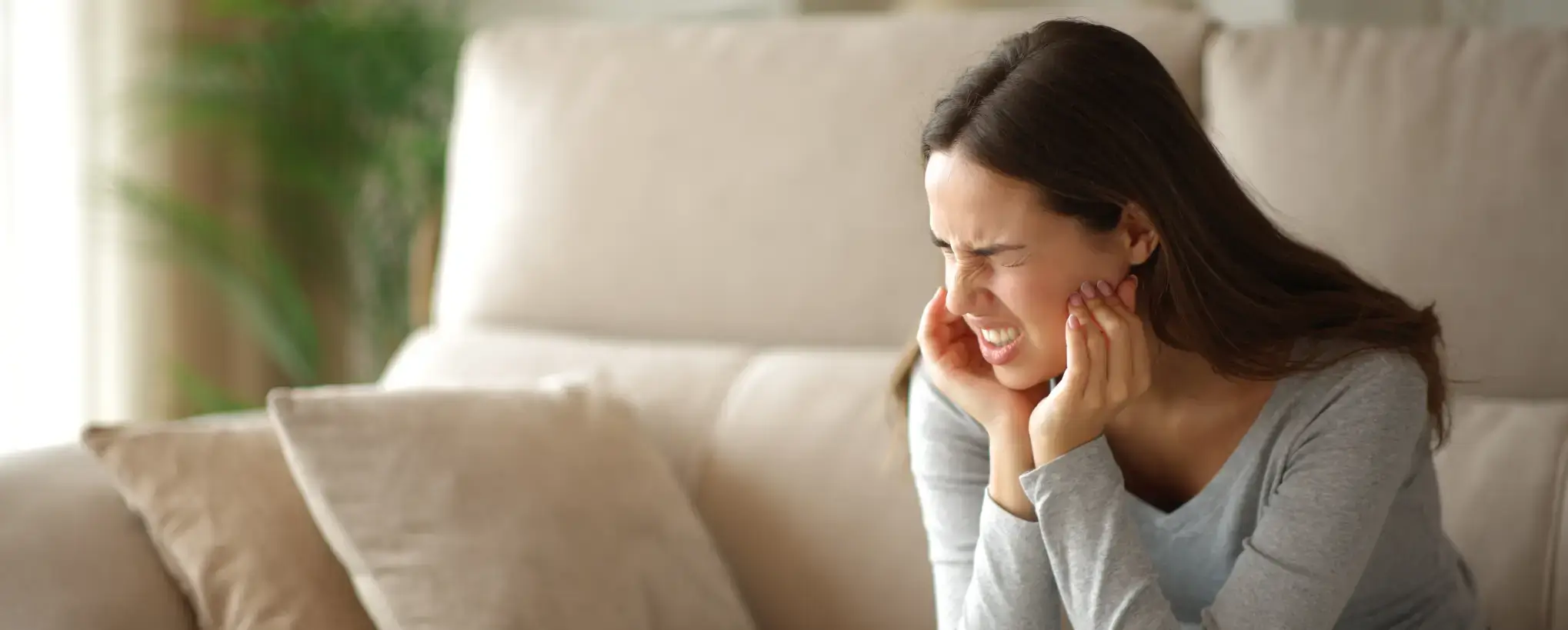 Woman sitting on a beige couch, grimacing in pain while holding her jaw with both hands.