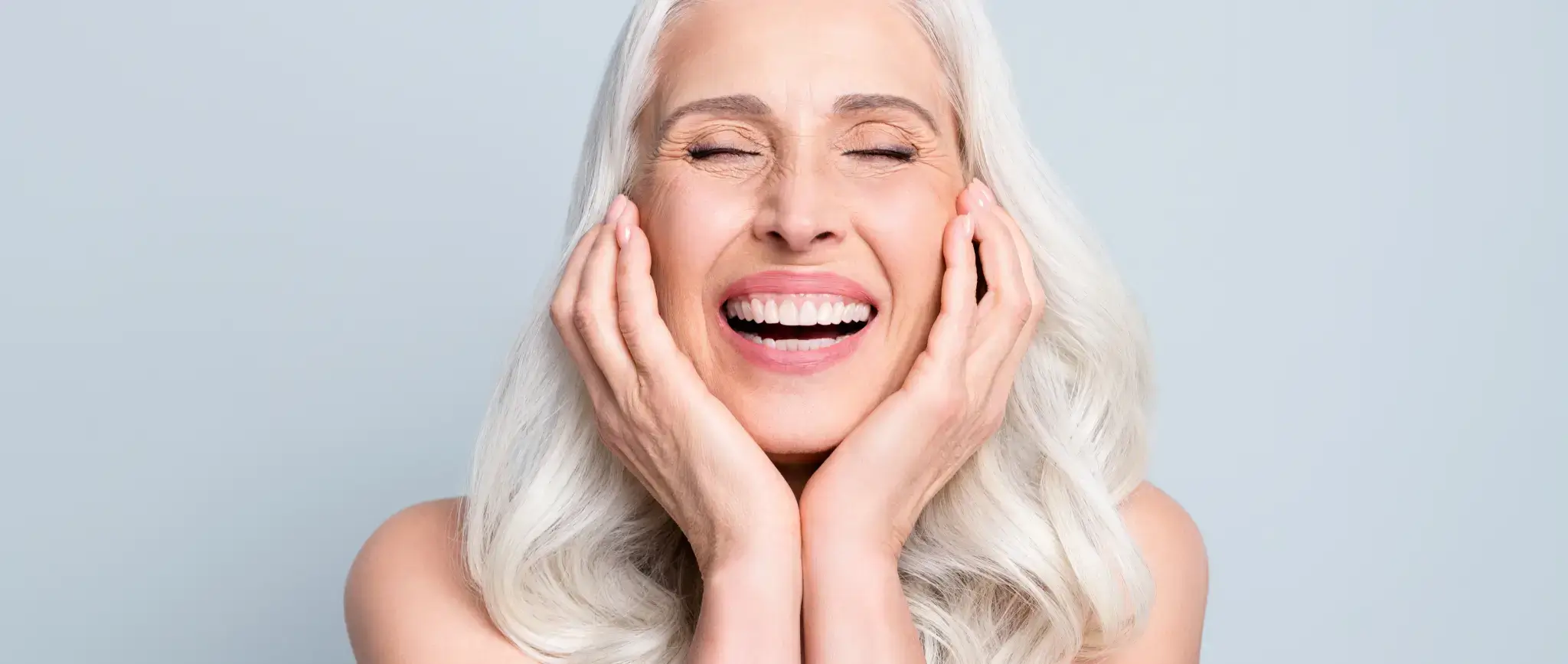 Elderly woman with long white hair smiling joyfully with eyes closed and hands on her cheeks against a light gray background.