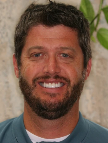 Smiling man with short dark hair and beard wearing a teal shirt in front of a leafy background.