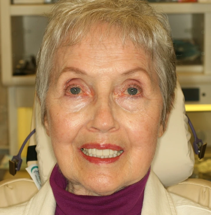 Close-up of an elderly woman with short gray hair, wearing a purple turtleneck and a light-colored jacket, smiling while seated in a chair.